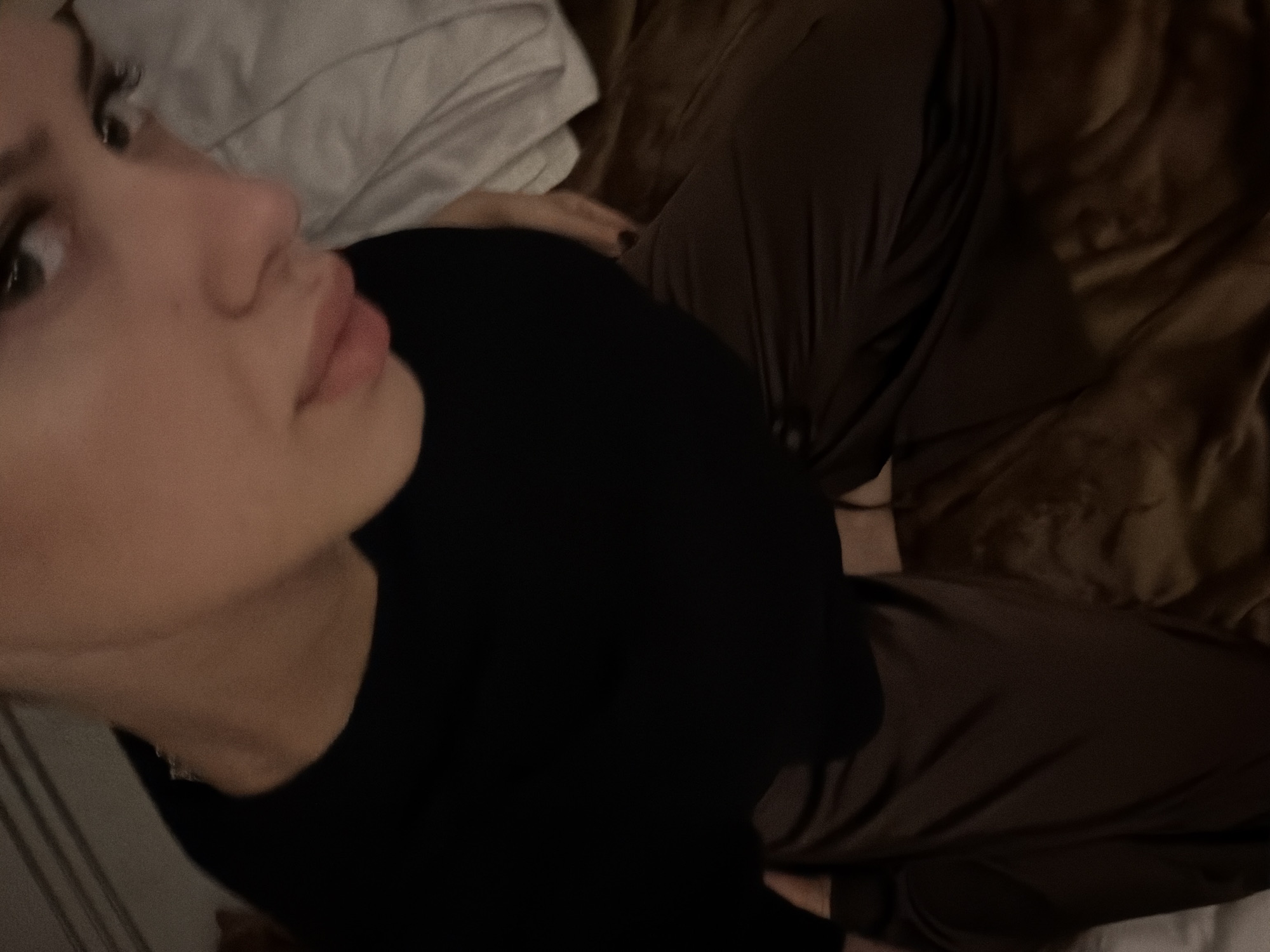 The image shows a close-up, high-angle shot of a person in a dark room, likely a bedroom. The person is wearing a black top and dark brown, shiny pants. Their face is partially visible, with their eyes looking towards the camera. The room appears to be dimly lit, with soft lighting that creates shadows and highlights on the person's clothing and skin. There is no indication of the person's location within the city of Đà Nẵng, Vietnam, or any other specific location. The scene is static, with no discernible action or interaction taking place. The focus is on the person's attire and the texture of the fabrics.