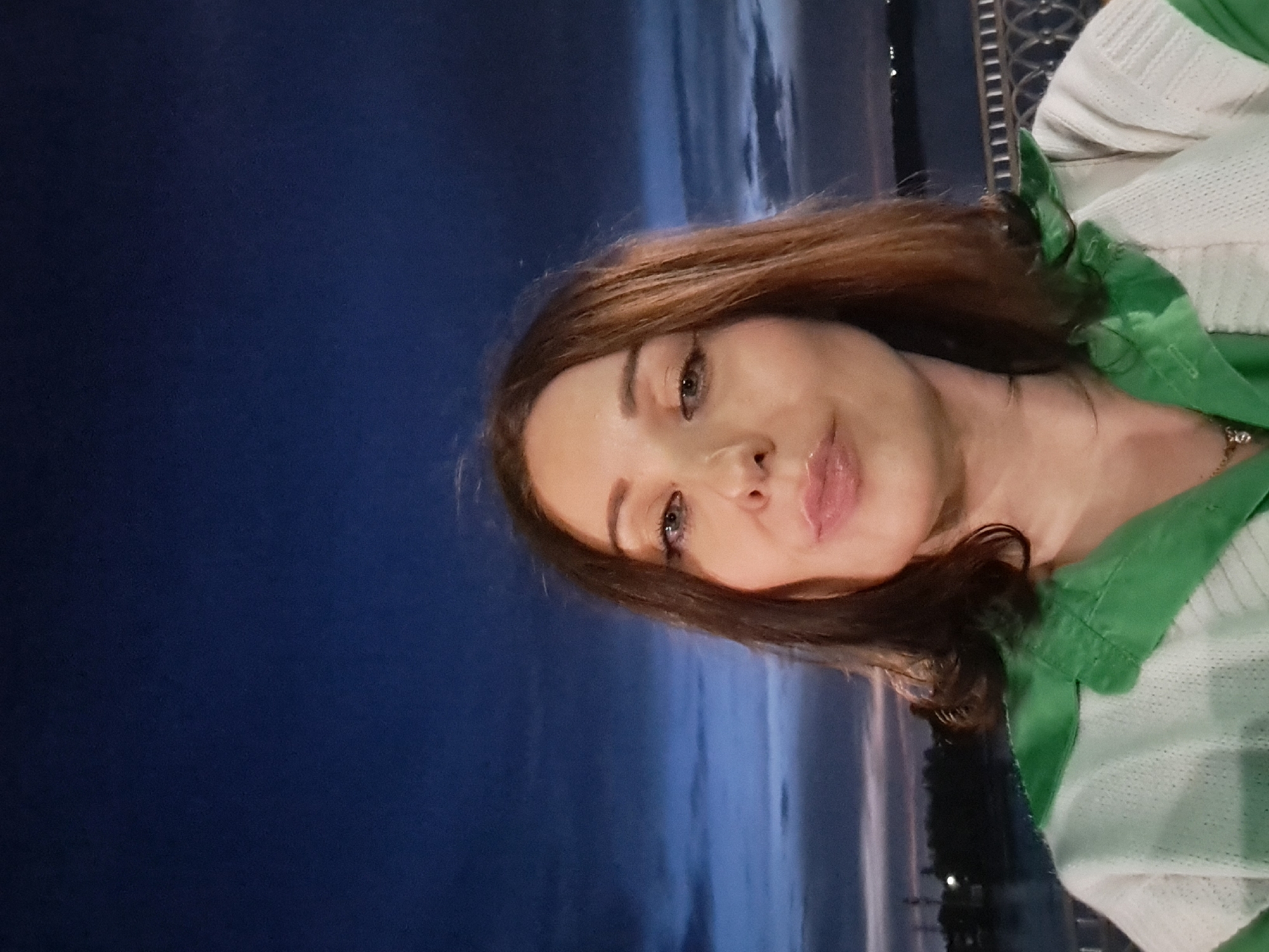 A woman is taking a selfie outdoors at dusk. The sky is a deep blue with some wispy clouds and hints of orange on the horizon. The woman has shoulder-length brown hair and is wearing a light-colored shirt with a green collar. She is looking directly at the camera with her lips slightly parted. In the background, a body of water is visible with some lights reflecting on it, suggesting a city or populated area across the water. A decorative metal railing is visible to the right of the woman. The specific location in Irkutsk, Russia cannot be determined from the image alone.