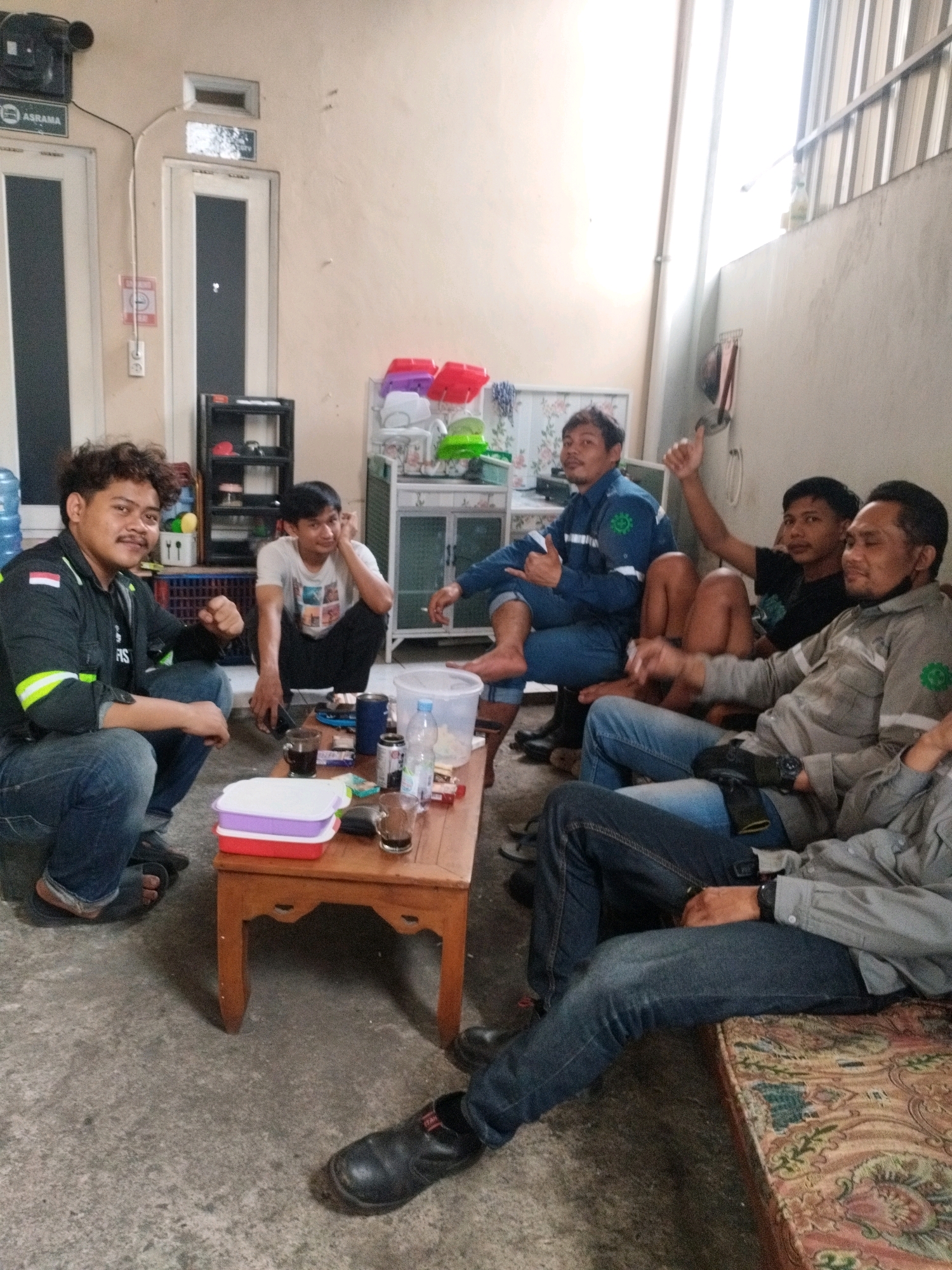 In Selomerto, Indonesia, a group of six men are gathered in an outdoor, covered area, possibly a workshop or a communal space. The men are seated in a relaxed manner, some on the floor and others on a low, cushioned bench. A wooden table in the foreground holds drinks and food containers. The environment appears to be informal, with bare concrete flooring and a wall constructed of light-colored plaster. A shelf unit with various items is visible against the wall behind the men. The men's attire suggests a work or casual context, with some wearing uniforms. The lighting is bright, indicating daylight, possibly filtered through a translucent roof.