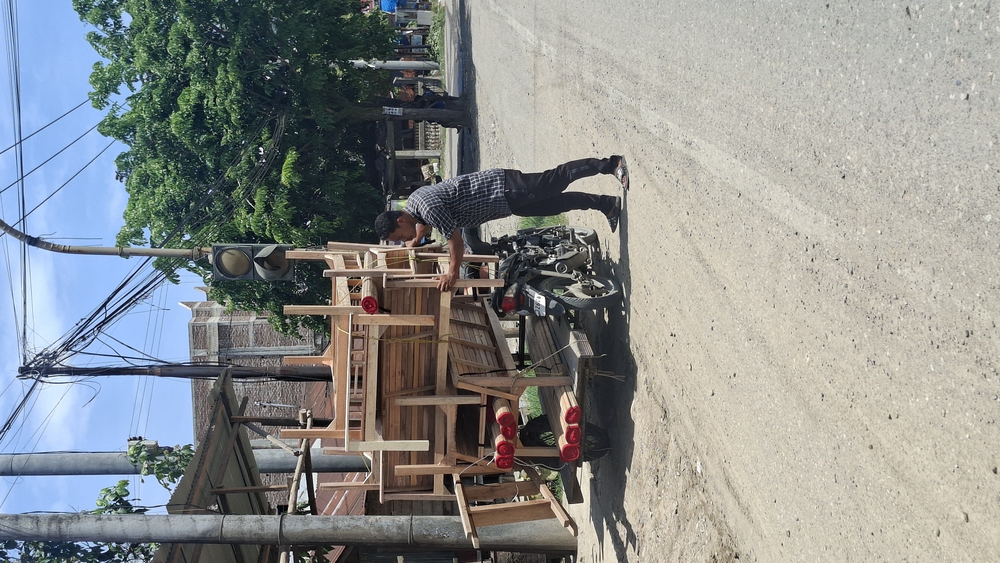 In Keudebayu, Indonesia, a man stands on a dirt road, pushing a trailer loaded with wooden furniture. The trailer is pulled by a motorcycle, which is parked behind it. The furniture, which appears to be newly made, is stacked high on the trailer. The man is wearing a plaid shirt and dark pants. The scene is set against a backdrop of trees and a clear blue sky. A traffic light is visible in the distance, suggesting that the location is near an intersection. The overall impression is one of a rural or semi-rural setting, possibly a market or a workshop area.