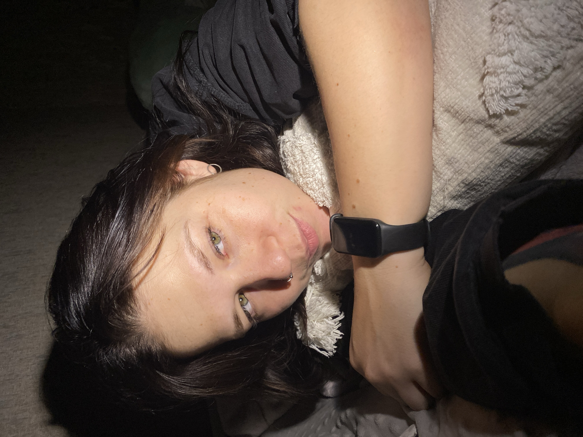 A close-up, overhead shot captures a young woman lying down in a dimly lit indoor setting in Chaikovsky, Russia. She has dark, shoulder-length hair and distinctive light-colored eyes, which appear green or hazel, looking directly at the camera. A septum piercing is visible on her nose. She is wearing a dark, possibly black, long-sleeved top or jacket, and a black fitness tracker or smartwatch is prominent on her left wrist.
Her head rests near a light-colored, textured blanket or pillow, suggesting a comfortable environment like a bed or sofa. The background is largely obscured by darkness, indicating low light conditions consistent with nighttime or a very dimly lit room. The lighting illuminates her face and arm from an angle, creating soft shadows. Her expression is calm and direct. No visible text is present in the image.