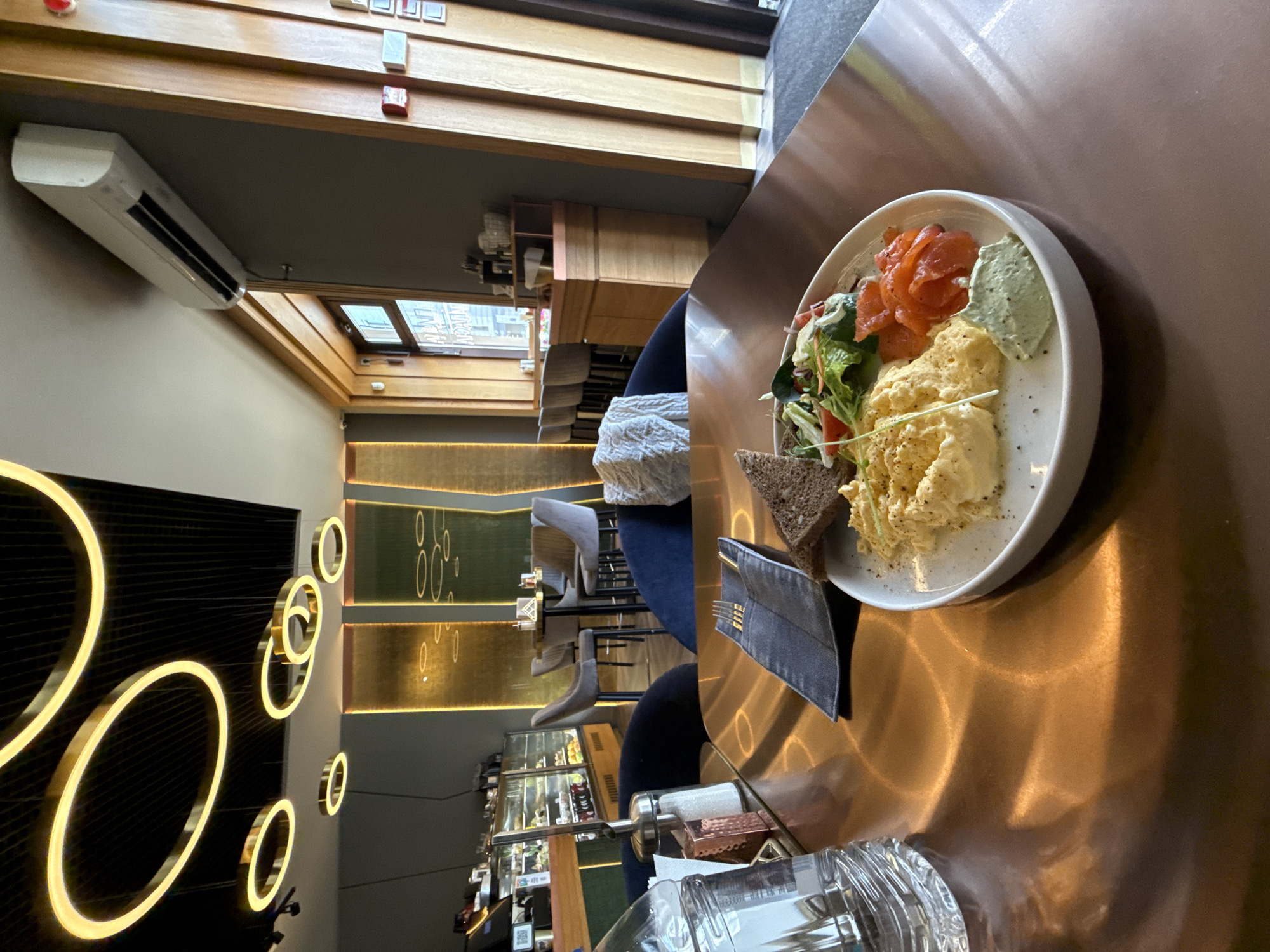 The image captures an overhead view of a meal served on a reflective, copper-toned table in what appears to be a modern cafe or restaurant. The main subject is a white plate featuring a portion of scrambled eggs, several slices of smoked salmon, a small side salad with mixed greens and possibly cherry tomatoes, and a dollop of light green sauce or spread. A slice of dark bread accompanies the meal. Next to the plate are a dark napkin holding cutlery, which partially reveals the text "BEE," a clear glass of water, and salt and pepper shakers in a caddy.
The setting is an indoor space with warm and contemporary aesthetics. In the background, decorative circular neon or LED lights of varying sizes illuminate a dark wall, casting soft glows. Wooden slatted panels adorn the ceiling and upper walls. An air conditioning unit is visible on an upper wall. Through a window or skylight, part of a building exterior with the text "LIV" on a frame can be seen, indicating daytime. The lower background features a bar or counter area with display cases, possibly showcasing pastries or desserts. Empty modern chairs and bar stools suggest a quiet period or ample seating. The overall ambiance is chic and inviting, likely set during the day given the natural light. A QR code is visible but unreadable on a small sign near the bar.