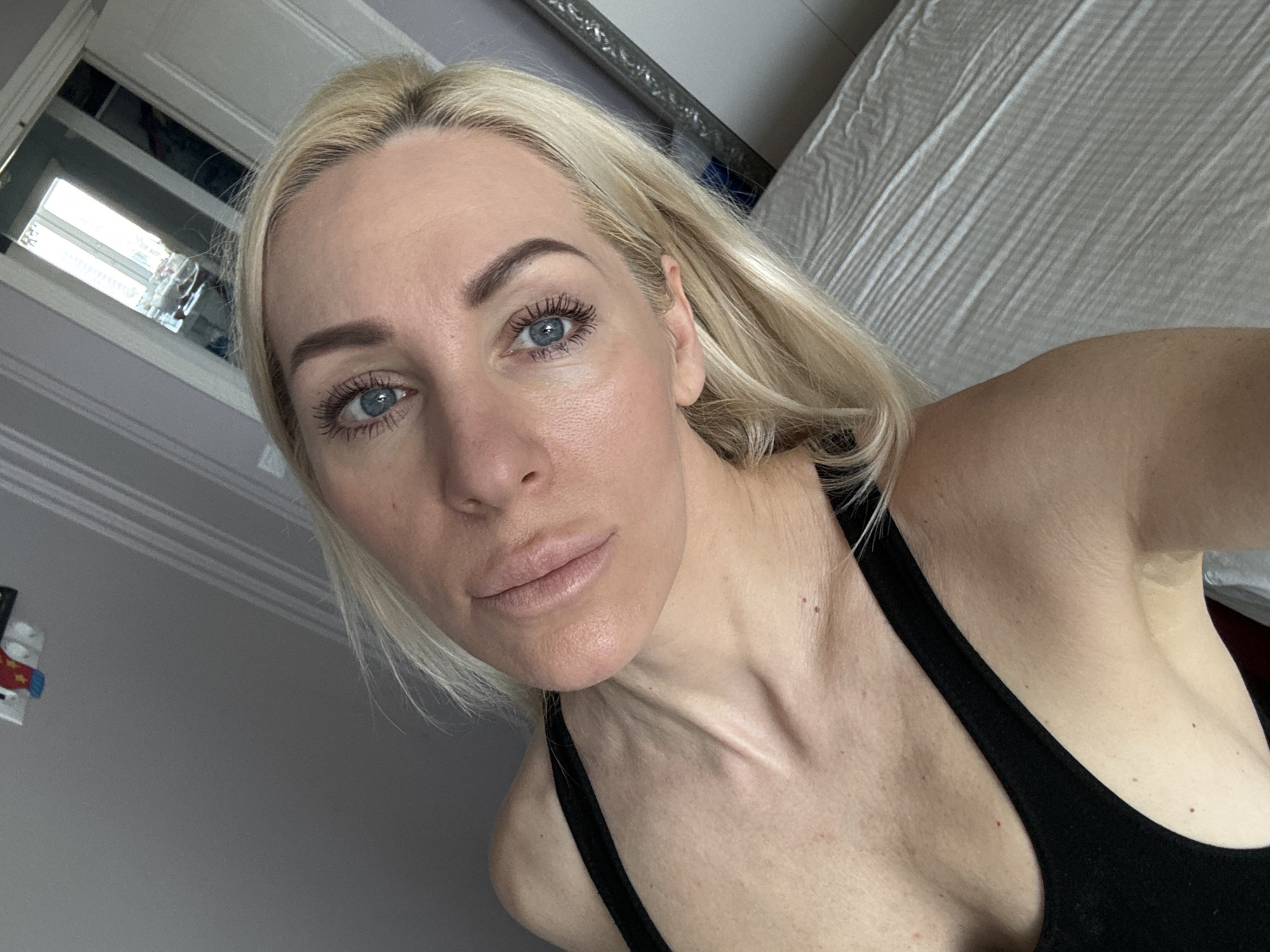 A close-up selfie shows a fair-skinned woman with blonde hair and blue eyes wearing a black tank top. Her hair is parted on the side. She is looking directly at the camera. The background includes a grey wall, a white window frame, a window with visible outdoor elements, and the edge of a white bed or linens. The location is not discernible from the provided image.
