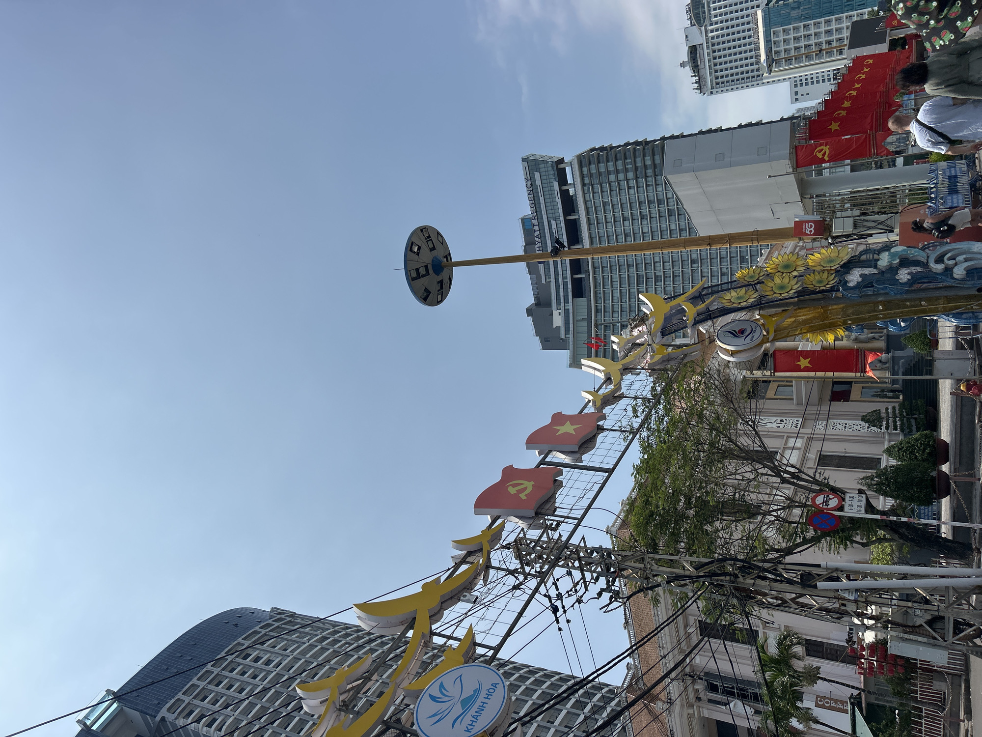 The image, taken in Nha Trang, Vietnam, captures a vibrant daytime street scene under a clear blue sky. Dominating the foreground are elaborate festive decorations, including stylized yellow elements and red banner-like shapes featuring the yellow star of the Vietnamese flag and the hammer-and-sickle emblem of the Communist Party of Vietnam. These are mounted on a metal framework, alongside a prominent circular sign reading "KHÁNH HÒA" with a bird/wave logo. Numerous tangled black overhead utility wires are also visible.
In the midground, a tall pole with a unique disc-like structure at its apex stands out. The background features a blend of modern and traditional architecture. A towering high-rise building prominently displays "HYATT REGENCY," while adjacent structures show rows of smaller red flags, some with the yellow star and others with the hammer and sickle, suggesting a patriotic or celebratory occasion. Further along the street are older-style buildings with ornate facades and green trees.
People are seen walking in the distance, engaging with the decorated urban environment. Visible text includes "KHÁNH HÒA," "HYATT REGENCY," "5G" on a red box, and "COFFE" on a sign. Traffic signs indicating "No Entry" and parking restrictions with specific times ("6:30-7:30" and "16:30-18:30") are also present. The overall scene conveys a lively and festive atmosphere in a bustling Vietnamese city.