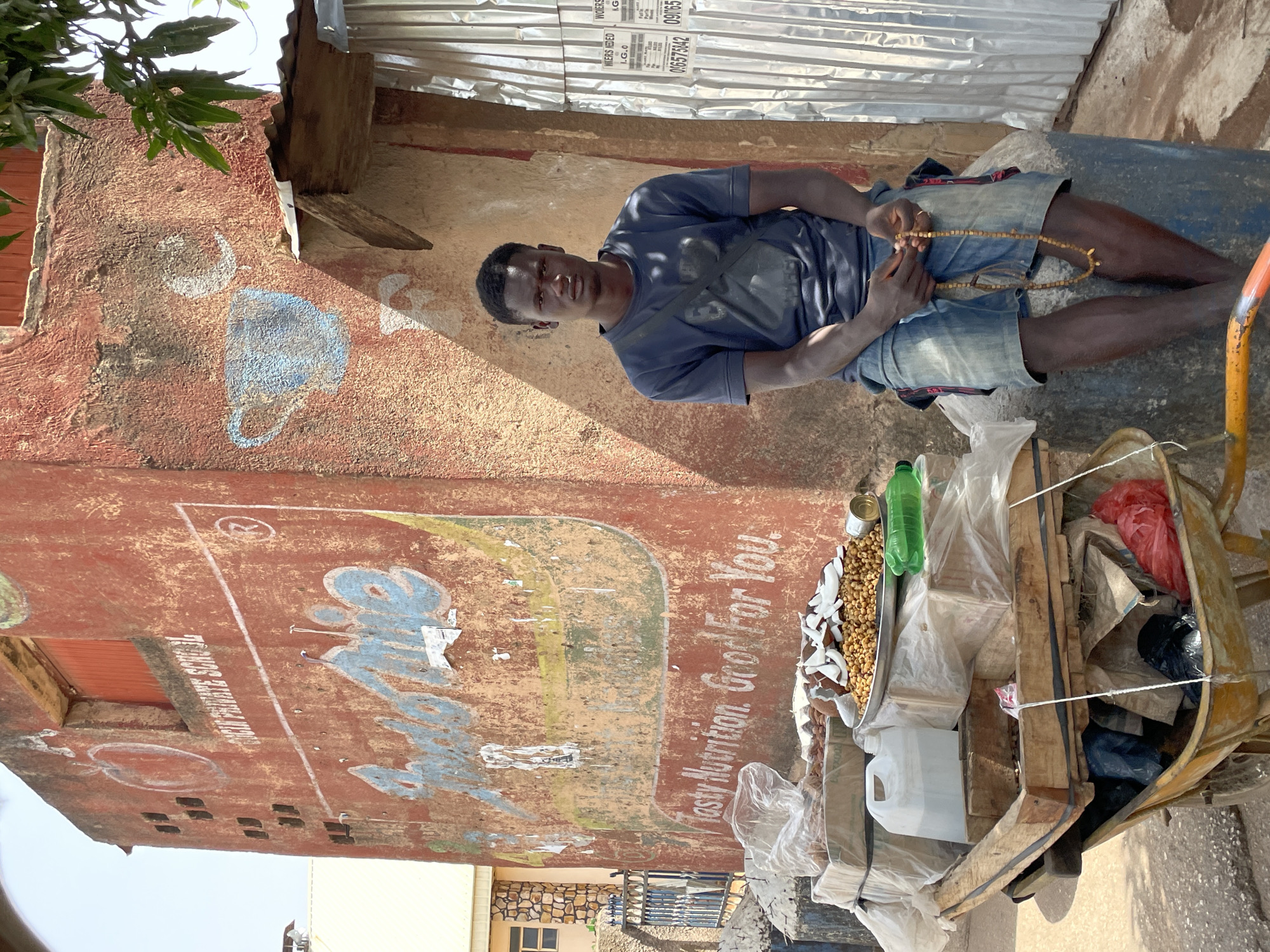 A man is sitting on a blue barrel next to a cart filled with goods in Jos, Nigeria. The man, wearing a blue shirt and jean shorts, is holding a string of beads. The cart contains various items, including coconuts and what appears to be nuts, in addition to plastic bags, bottles, and containers. The cart is a wooden structure with yellow and orange metal components. Behind the cart and the man is a building with faded advertising paint on its reddish-brown exterior, including the text "Tasty Nutrition. Good For You". The area appears to be an outdoor market or commercial street, with another building visible in the distance. The background includes a corrugated metal roof structure.