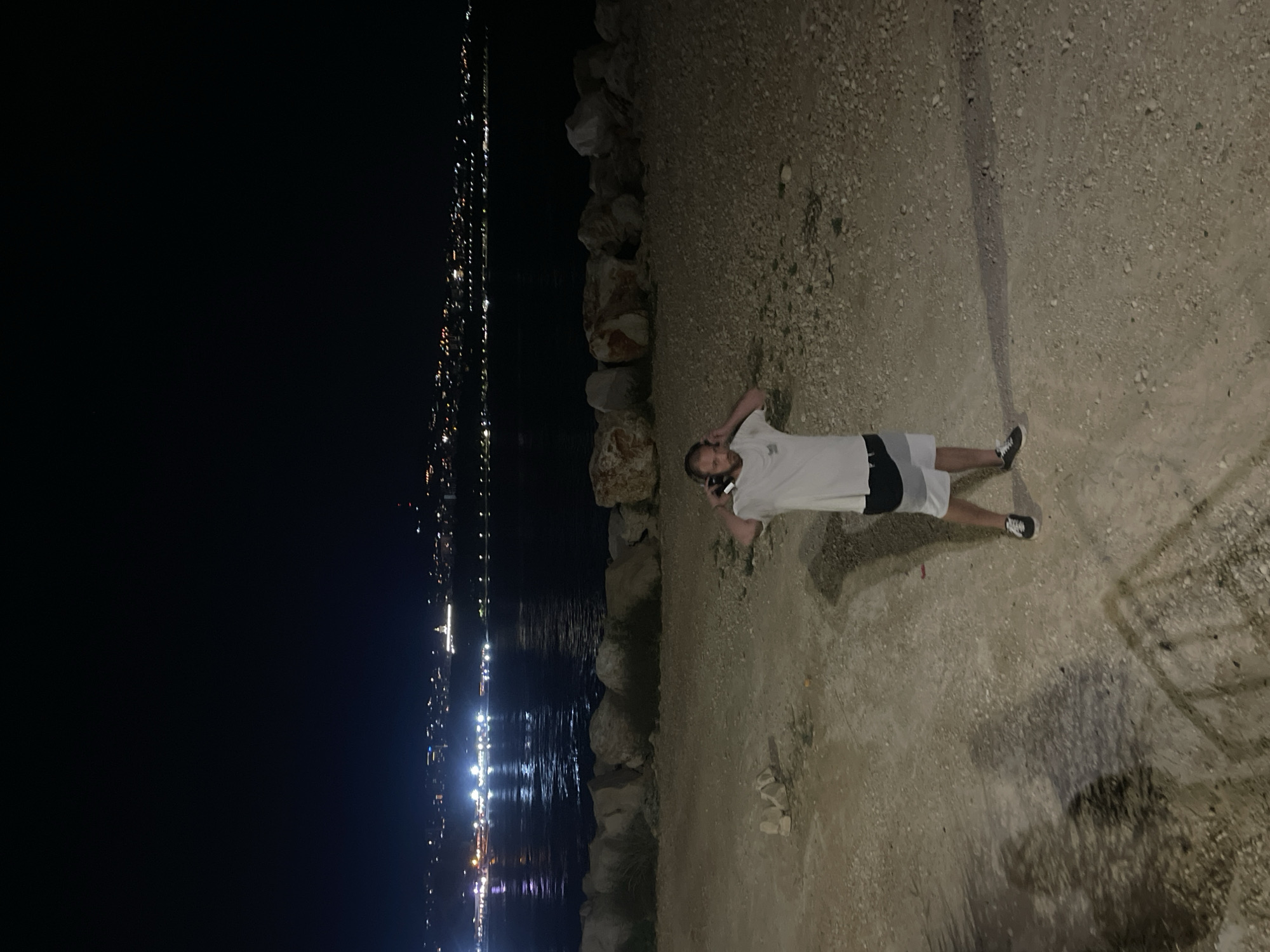 A full-body, medium shot captures a man standing on a rocky shore at night, facing the camera. He is wearing a white t-shirt with a graphic design, white and black patterned shorts, and dark sneakers. The man has headphones on and appears to be holding his phone to his ear, as if on a call. The background features the calm, dark water of the sea, with a distant city skyline illuminated by numerous lights stretching along the coast. The shoreline in the foreground is composed of sand and scattered rocks. The scene is set at night, with artificial lights from the city and what appears to be a bright light on a structure in the distance reflecting on the water. The location is the Varna Sea Garden, specifically a section of the rocky shoreline with a view of the city lights.