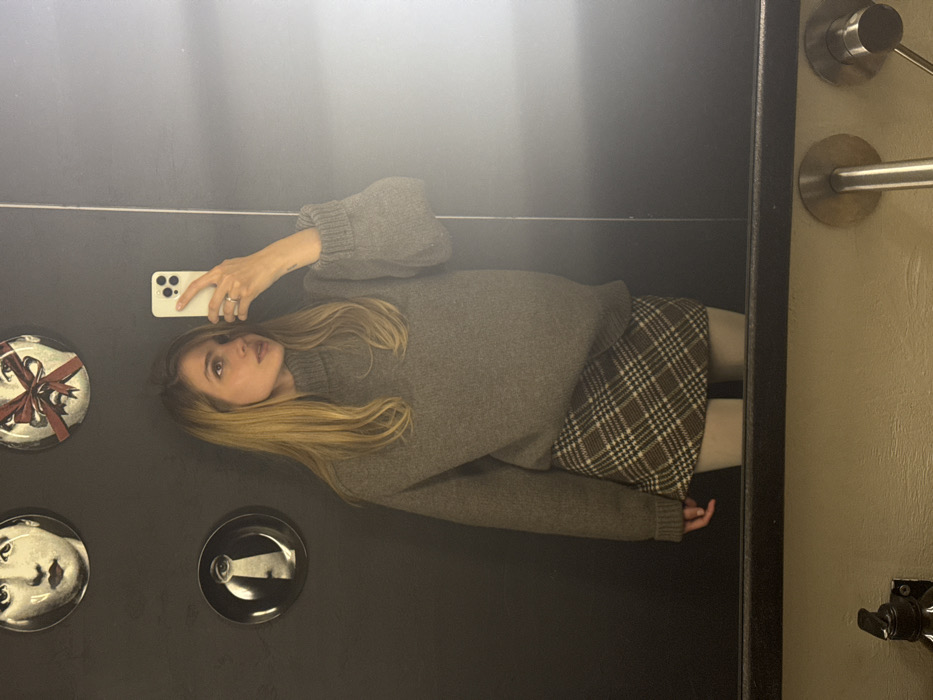 A woman with blonde hair wearing a gray turtleneck sweater and a plaid skirt takes a selfie in a mirror. Her phone is visible in her left hand. Decorative plates adorn the wall behind her. The mirror is framed by a black border, and a section of a beige wall with metallic hardware is visible on the right side of the frame.