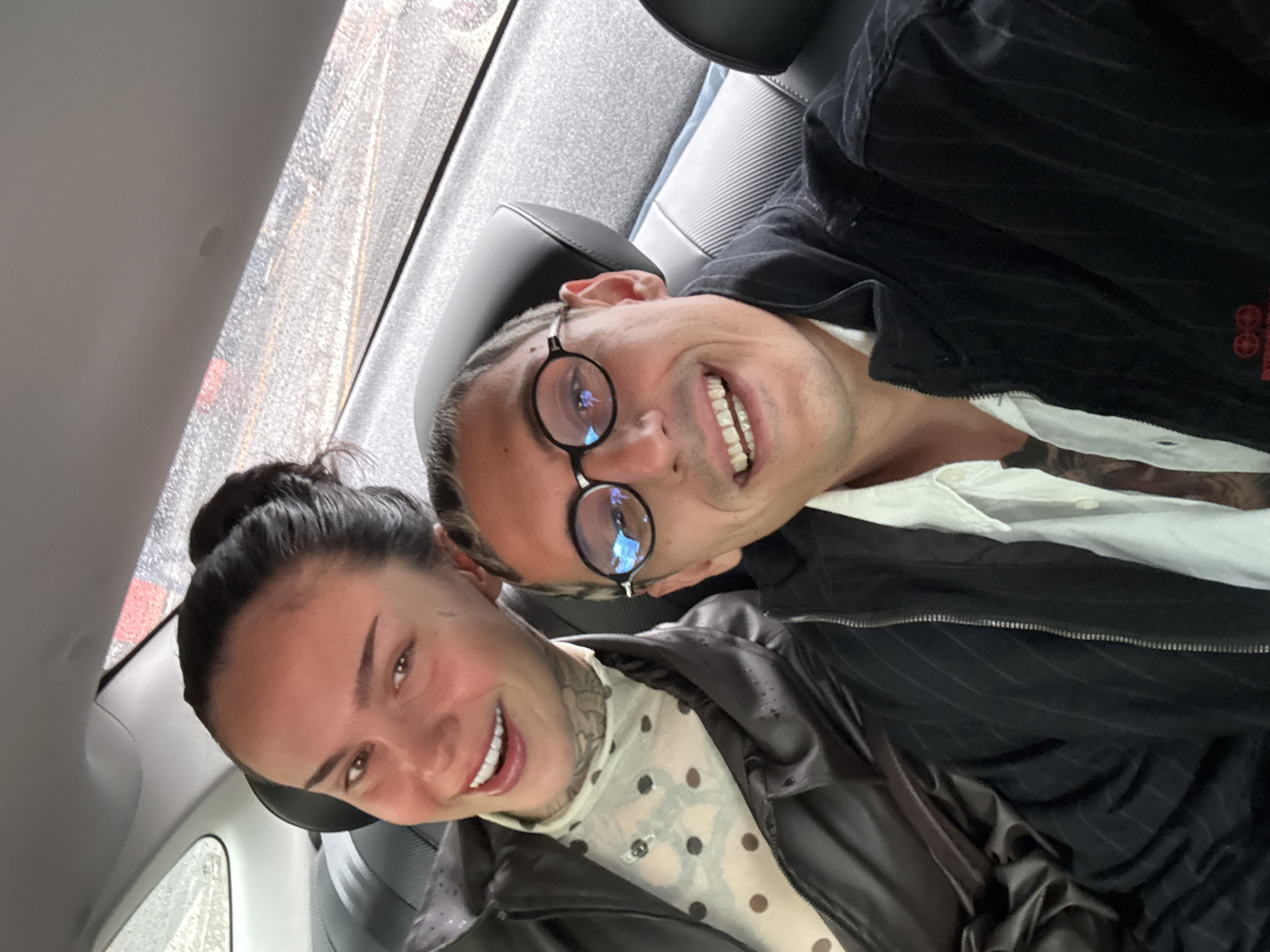 Two people are sitting in the back of a car. The woman has her hair up in a bun and is wearing a brown leather jacket over a polka dot top. The man is wearing glasses and a dark jacket over a white shirt. Rain is visible on the car window. It is not possible to determine the location of this image as Seoul, South Korea.