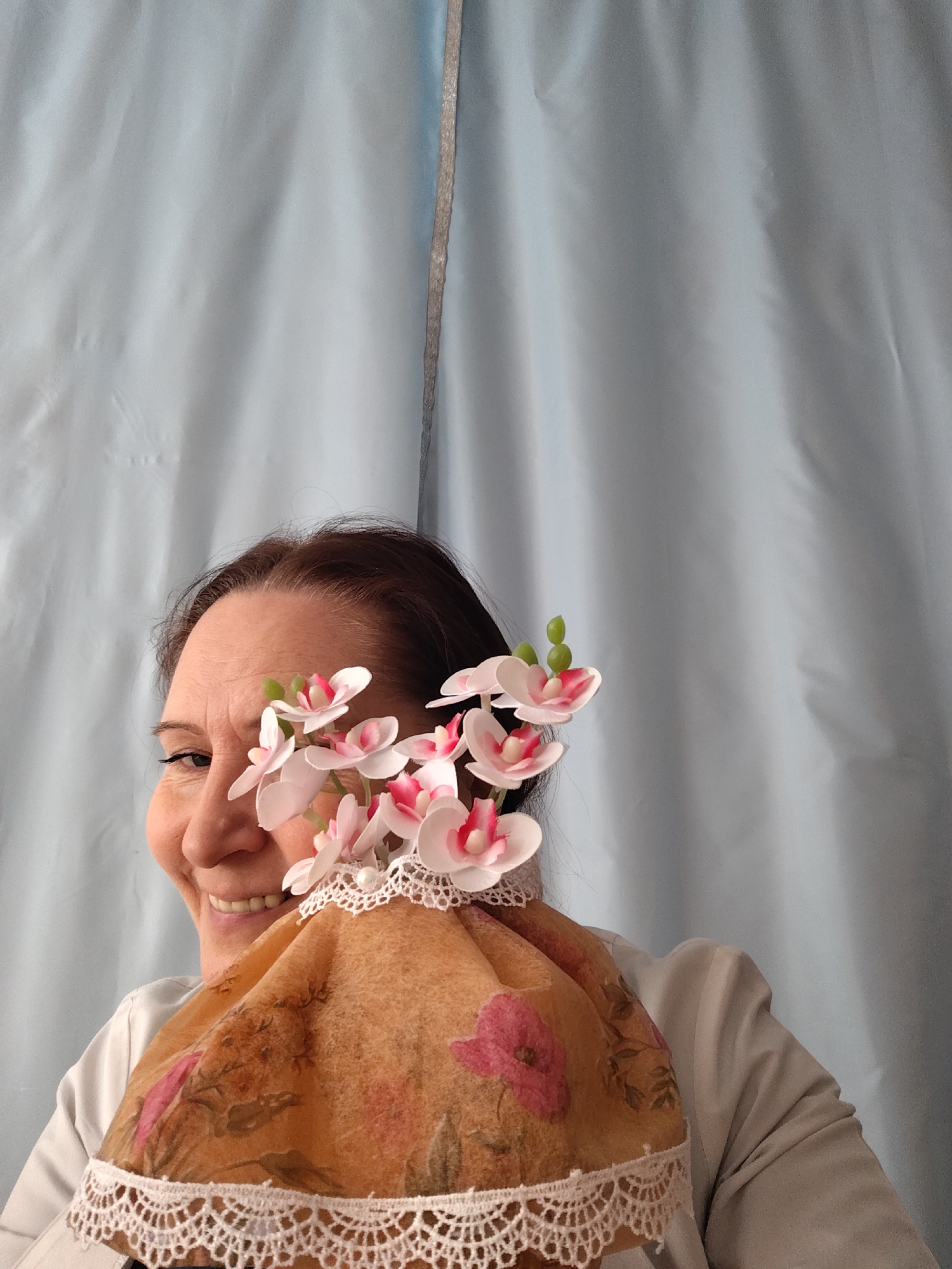 A woman is partially visible, smiling and holding a brown, poppy-patterned cloth with a white lace border and a cluster of pink and white flowers that partially obscure her face. She is wearing a white blazer-style garment. The background consists of a light blue, somewhat creased curtain. There is no information in the image to confirm a location in Sarapul, Russia.