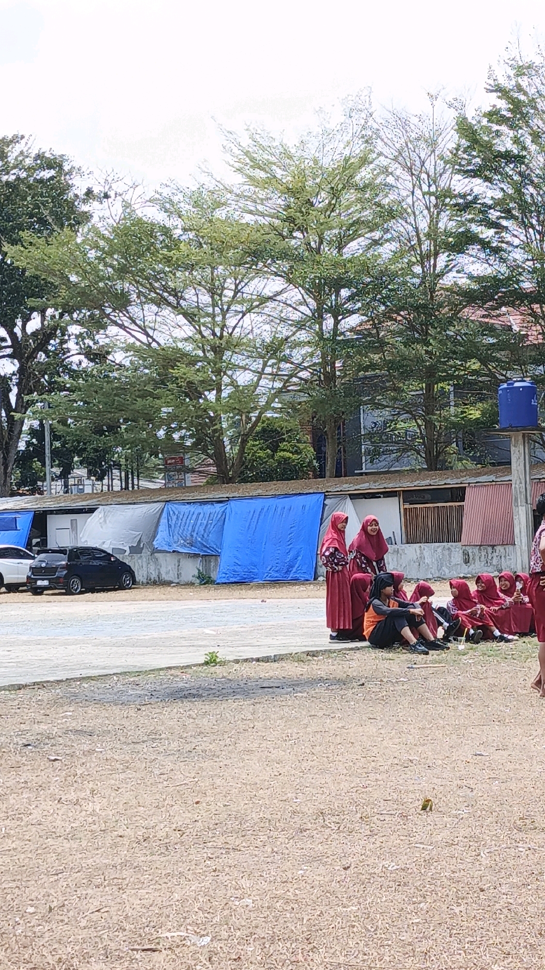 In Cikembulan, Indonesia, a group of young people, predominantly female, are gathered outdoors on a dry, dusty ground. Several are seated, with some wearing headscarves and maroon clothing. One individual in an orange shirt and black pants sits on the ground, facing left, with their legs crossed and hands resting on their knees. Others in the background are standing, also wearing headscarves and maroon attire. A few cars are parked to the left of the scene, near a row of structures covered with blue and gray tarps. Trees with sparse green foliage are visible behind the structures, and a blue water tank stands on a pole to the right. The sky is overcast with a bright, pale tone.