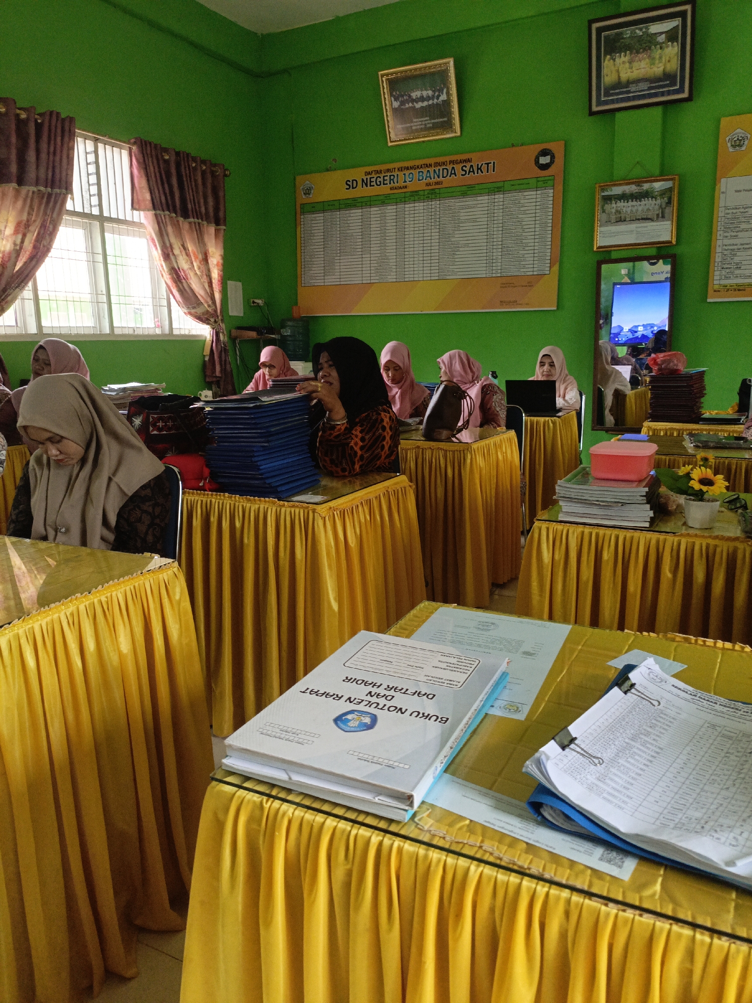In a brightly lit room with emerald green walls, several women wearing hijabs are seated at tables draped with vibrant yellow cloths. The room appears to be a meeting or classroom setting. A large bulletin board displaying a chart with rows of names and numbers is mounted on the wall. Framed photographs of groups of people in uniform attire are also displayed on the walls. On the tables, stacks of blue folders, books, and papers are neatly arranged. One woman is actively engaged with a laptop, while another is looking down at a document. In the foreground, a book titled "BUKU NOTULEN RAPAT DAN DAFTAR HADIR" (Meeting Minutes and Attendance Book) is prominently placed on a table. The overall impression is one of an organized gathering or event.
The setting of this image is a room within "SD Negeri 19 Banda Sakti" (Public Elementary School 19 Banda Sakti), located in the city of Lhokseumawe, Indonesia.