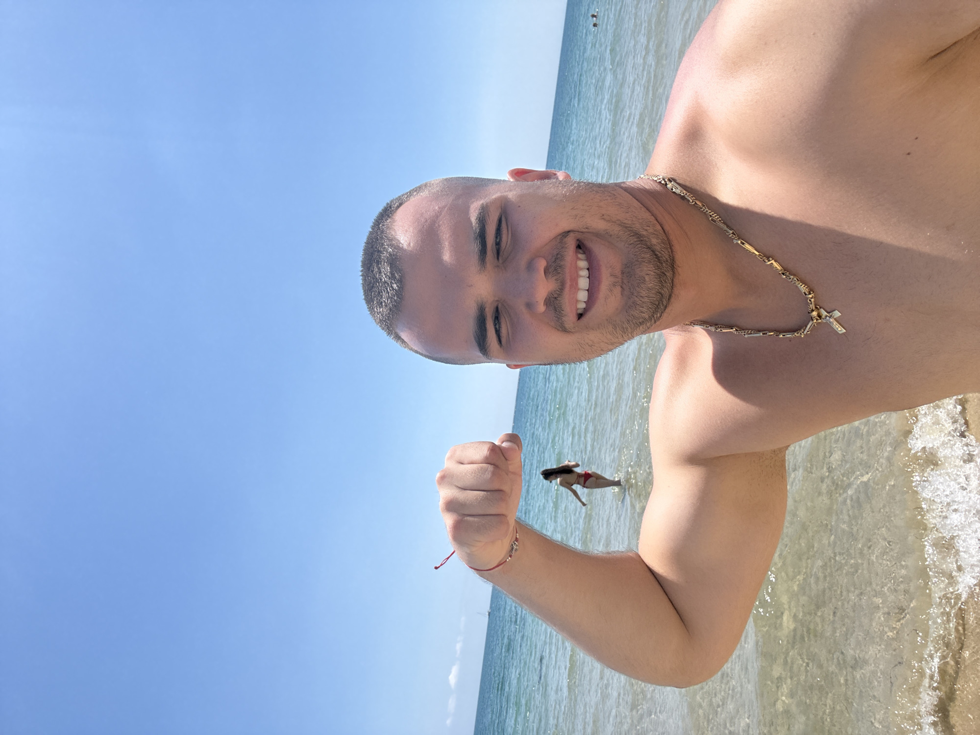 A man with a shaved head, a gold chain, and a red string bracelet on his left arm is flexing his bicep at the camera. He is smiling. He is standing in the ocean with clear water. In the background, a woman in a red bikini is standing in the water. The sky is clear blue and sunny. The location is the beach.