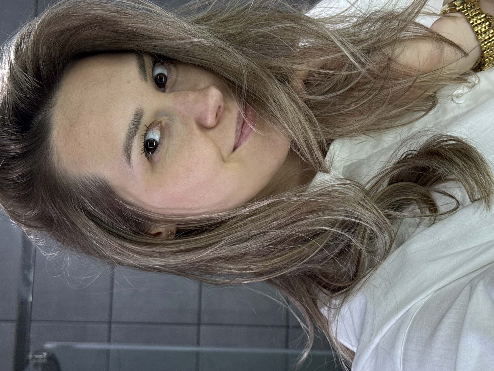 A young woman with light brown hair, styled with soft waves, is posing for a selfie. She is wearing a white shirt and a gold watch is visible on her wrist. The background appears to be a tiled wall, possibly in a bathroom. There is no indication of her location being in Chaikovsky, Russia.