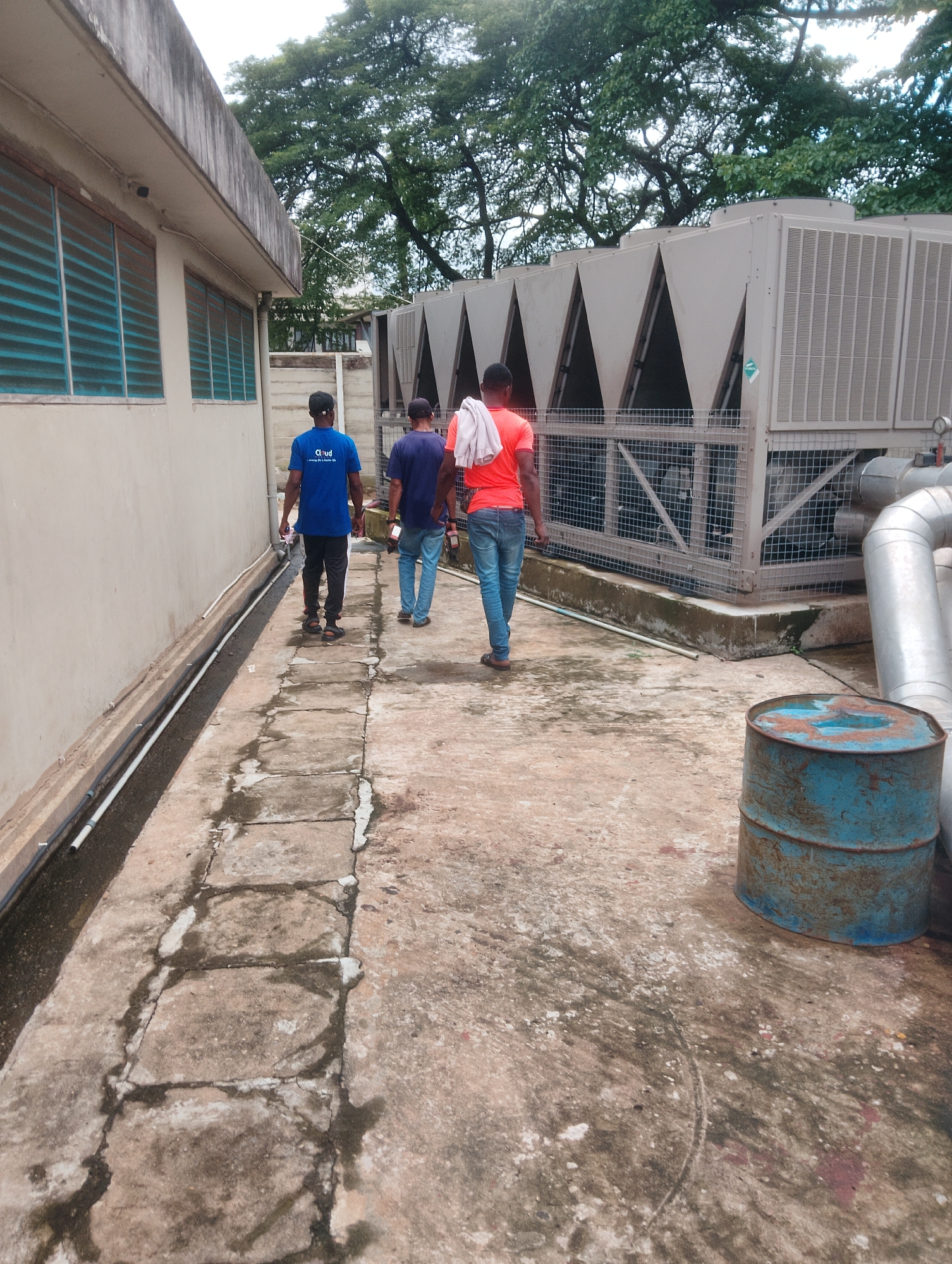 Three men walk away from the camera on a cracked concrete walkway next to a building. The man on the left wears a blue t-shirt with white text that reads "Cloud" and "Energy For A Better City". He also wears black pants and a black cap. The middle man wears a dark blue t-shirt and jeans. The man on the right wears an orange t-shirt with a towel draped over his shoulder and blue jeans. Behind the men is a large industrial air conditioning unit. To the right of the walkway, there is a rusty blue barrel and large metal pipes. The building to the left has horizontal blue-tinted windows. Trees are visible in the background. The ground is wet in some areas, suggesting recent rain or a damp environment. The sky is overcast, indicating a cloudy day. The location is identified as Onibudo, Nigeria. The activity appears to be a work-related inspection or movement between areas.