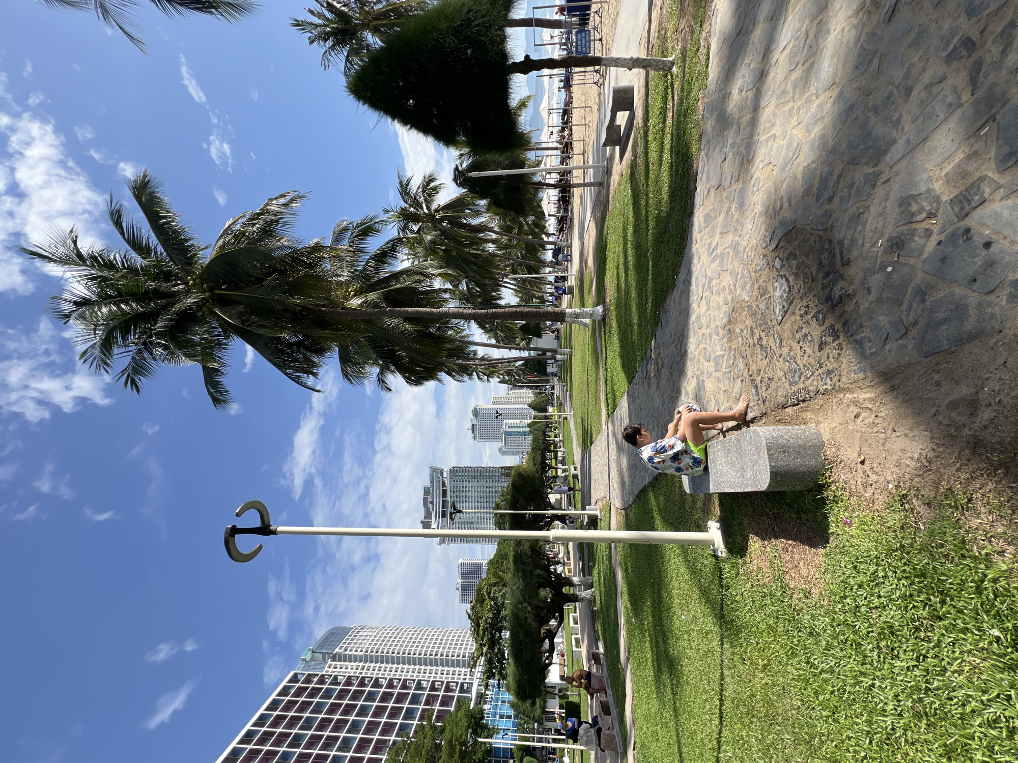 A young boy, wearing a colorful short-sleeved shirt and green shorts, sits on a concrete bench on a paved walkway adjacent to a manicured lawn. He is looking down at something in his hands. The walkway is lined with palm trees and other greenery. In the background, several tall, modern buildings are visible, with a blue sky and scattered white clouds overhead. The scene appears to be a public park or promenade in Nha Trang, Vietnam, likely along the beachfront, given the presence of palm trees and the implied proximity to a beach. Other people are visible in the distance, some sitting on benches and others walking. The overall atmosphere is bright and sunny, suggesting a warm day.