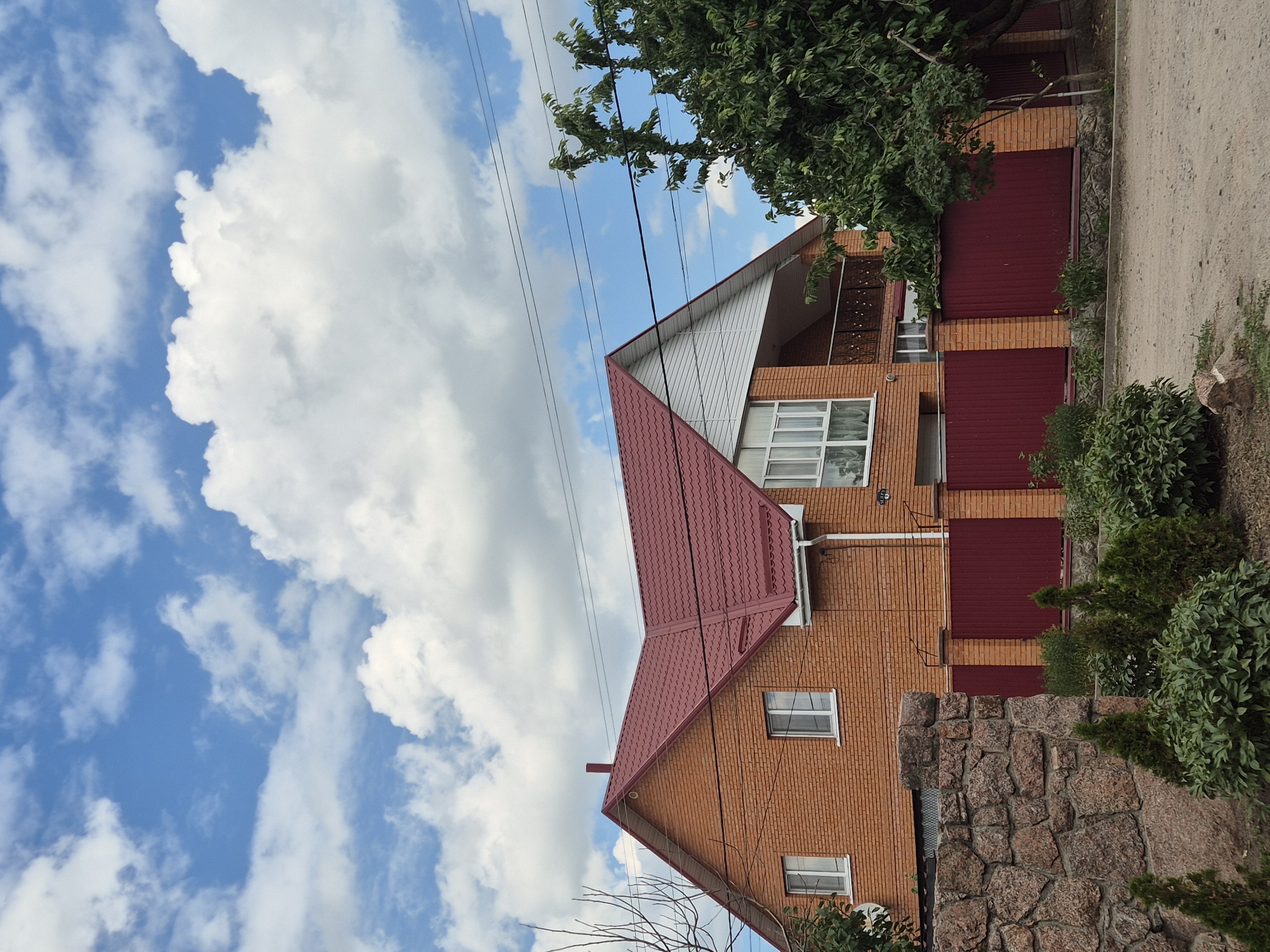 This is a vertical, eye-level medium shot of a two-story brick house with a red metal roof under a blue sky with scattered white clouds. The house appears to be under construction or recently built, with clean brickwork and modern white-framed windows. A dark red metal fence runs along the side of the property, separating it from a dirt road. Lush green bushes and trees are visible around the house and fence. Power lines stretch across the upper portion of the frame, and a portion of a stone wall is in the foreground. The image is well-lit, suggesting it was taken during the daytime, likely on a clear or partly cloudy day. The scene evokes a sense of suburban or rural domesticity. There is no visible text in the image. The location context provided is Sozonivka, Ukraine.