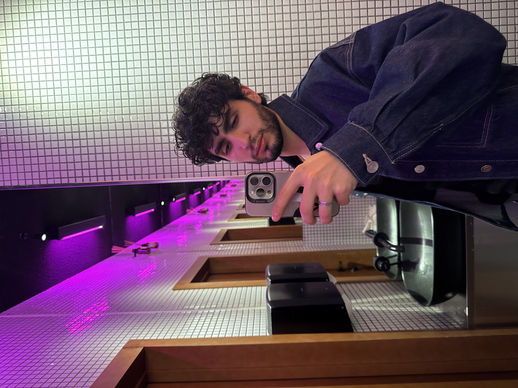 A man with dark, curly hair and a beard is taking a selfie with a smartphone in what appears to be a restaurant or bar. He is wearing a dark denim jacket. The background consists of a white tiled wall and a long counter illuminated with purple neon lights. The counter has built-in compartments with what appears to be plates or trays. The style of the establishment may be modern or minimalist. The location is Yerevan, Armenia.