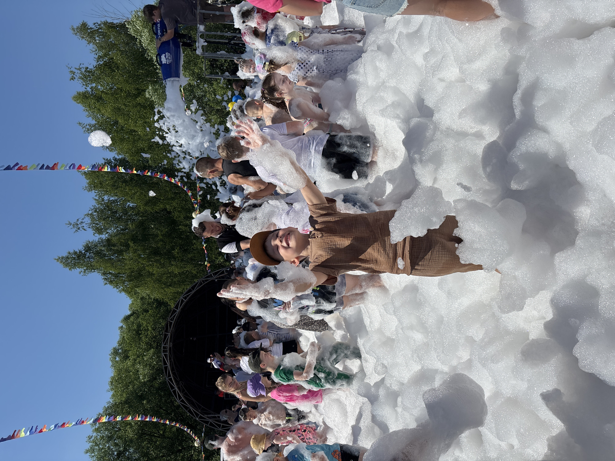 In a sun-drenched outdoor setting, a lively foam party is in full swing. A dense white foam covers the ground and envelops a crowd of people, mostly children and young adults, who are joyfully immersed in the frothy expanse. Many are seen splashing and playing in the foam, their faces alight with excitement and laughter. Some individuals are lying down, while others are standing or sitting amidst the bubbles.
In the background, lush green trees frame the scene against a clear blue sky, suggesting a warm and pleasant day. A stage structure is partially visible, hinting at a possible event or performance taking place. Colorful festive banners are strung across the area, adding to the celebratory atmosphere. The overall mood is one of uninhibited fun and delight, with people of all ages participating in the playful activity. One person in the foreground, wearing a brown top and a hat, looks up and smiles towards the camera, their hands raised and covered in foam. The abundance of foam creates a surreal and magical environment.