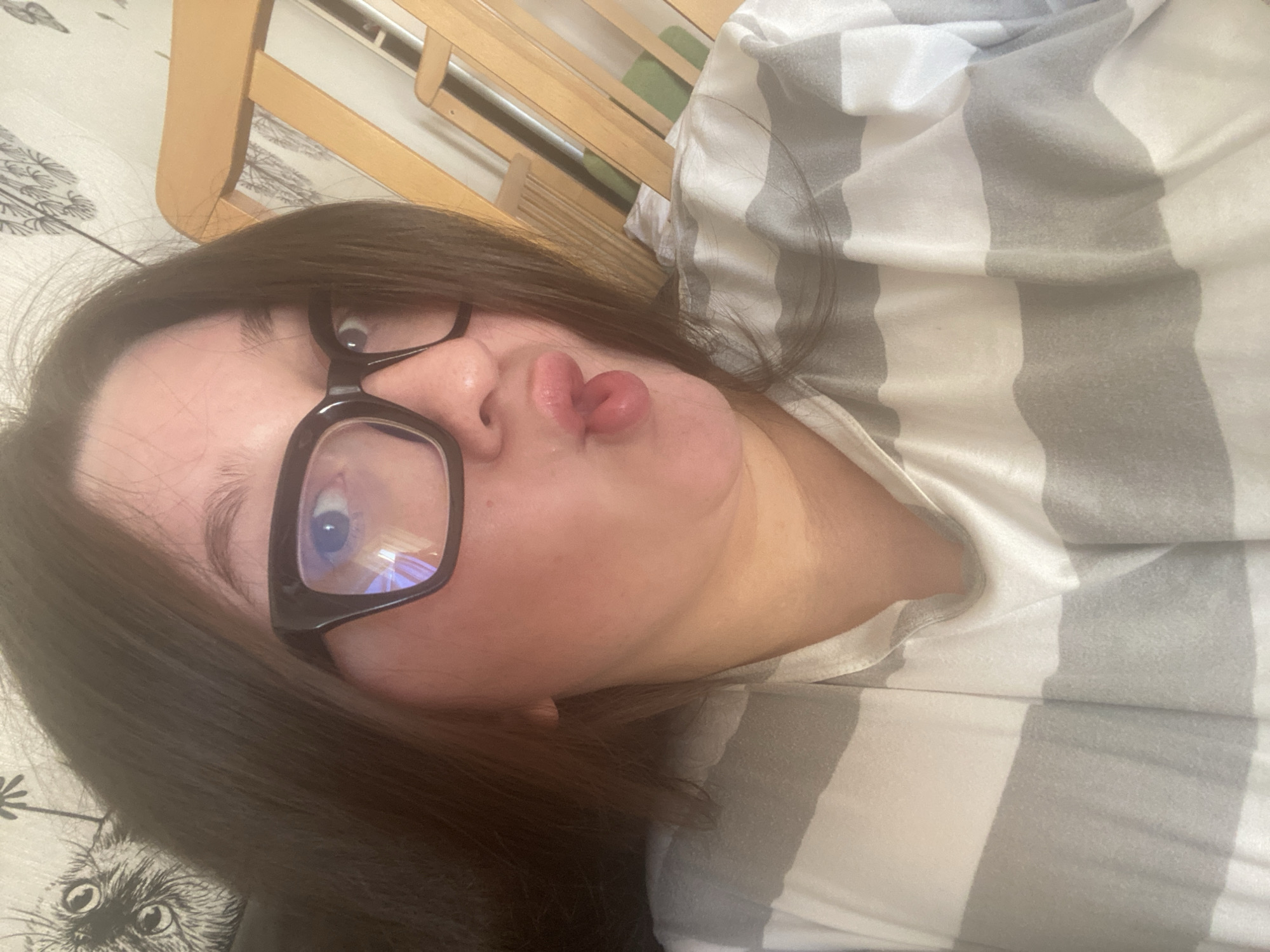 A female individual with dark brown hair and black-framed eyeglasses is depicted in a close-up, chest-up indoor shot. She is wearing a white and gray horizontally striped shirt. Her lips are puckered, forming a "duck face," with the tip of her tongue extended. Reflections are visible in the lenses of her eyeglasses. The background on the left features a dark, unidentifiable surface and a light-colored wallpaper with a repeating pattern of black, stylized animal heads, possibly cats or owls. On the right, a light-colored wooden structure with vertical slats, likely part of a bed frame or similar furniture, is visible against a plain light wall. A glimpse of a green object or fabric is discernible behind the wooden slats. The precise location within Chaikovsky, Russia, cannot be determined from the provided visual evidence.