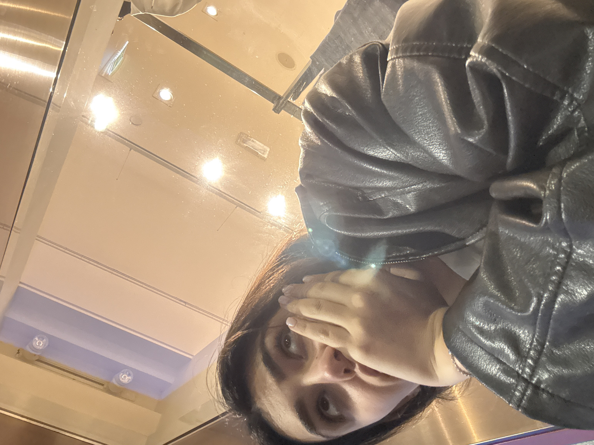 A person is reflected in a glossy surface, possibly a glass table or display case, with a light-colored ceiling and recessed lighting visible in the reflection. The person is wearing a dark, possibly leather, jacket. The individual's face is partially obscured by their hand. The location cannot be determined to be Milan, Italy based on the provided information.