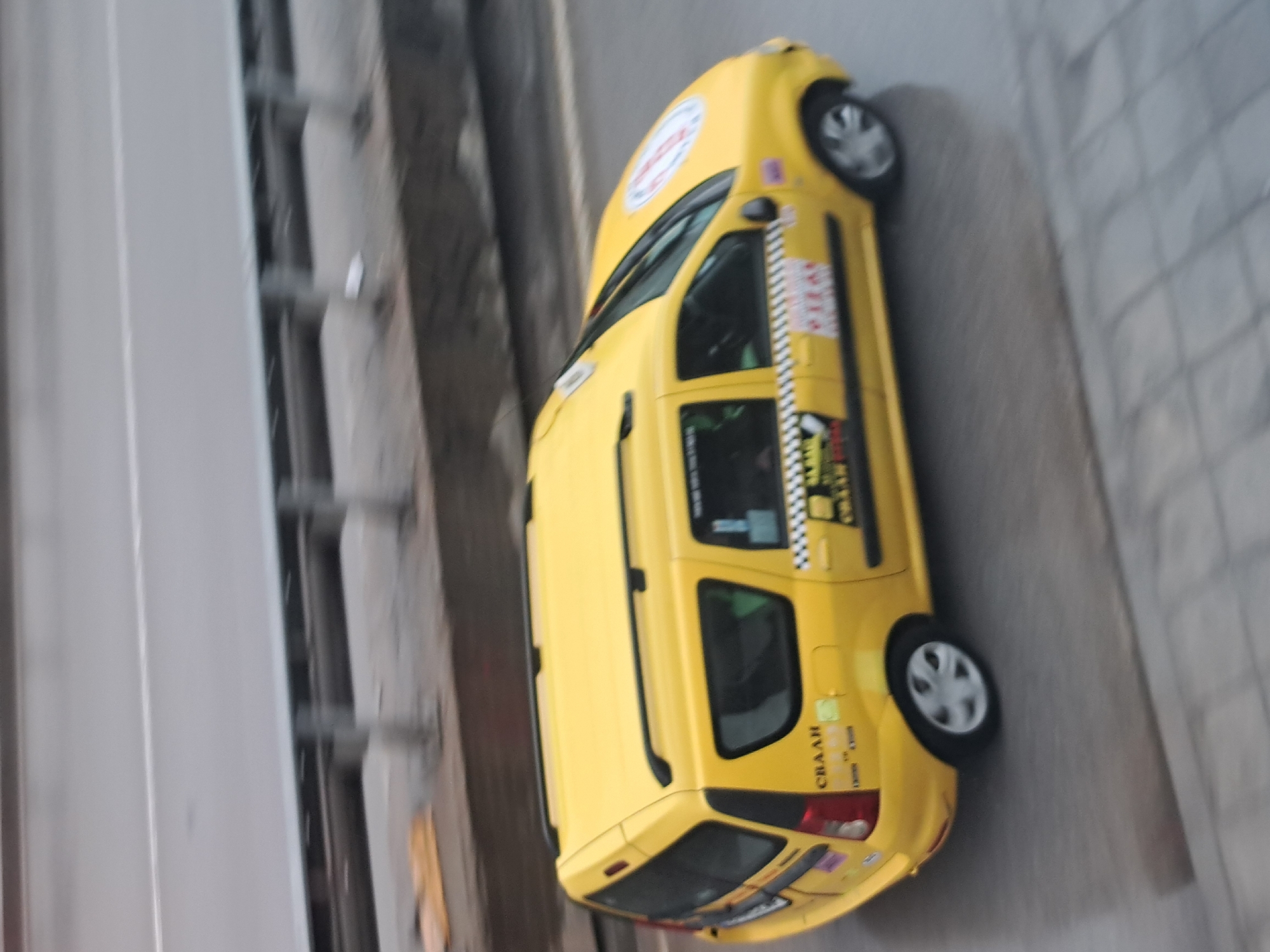 The image, taken from an elevated perspective, shows a bright yellow taxi driving or parked on a paved road under clear daylight. The car is a five-door station wagon, possibly a Dacia Logan MCV, with black roof rails.
On the roof, there is a white circular logo with red text, which appears to be "ТАХІ" (Cyrillic for Taxi). Along the sides of the car, specifically on the doors, there is a prominent black and white checkered stripe, typical of taxis, with red Cyrillic text reading "ТАХІ" above it. Below the rear side window, more Cyrillic text is visible, including "СВААН" and other smaller, less decipherable words. A similar white circular logo with red text is also visible on the front quarter panel or fender. The windows are tinted.
To the left of the car, a sturdy grey concrete structure runs parallel to the road, suggesting an overpass, bridge, or elevated roadway. On the right, the paved road features a section with a grid-like or tiled pattern, indicating a sidewalk or a different type of paving. The overall lighting suggests it is daytime with good visibility.