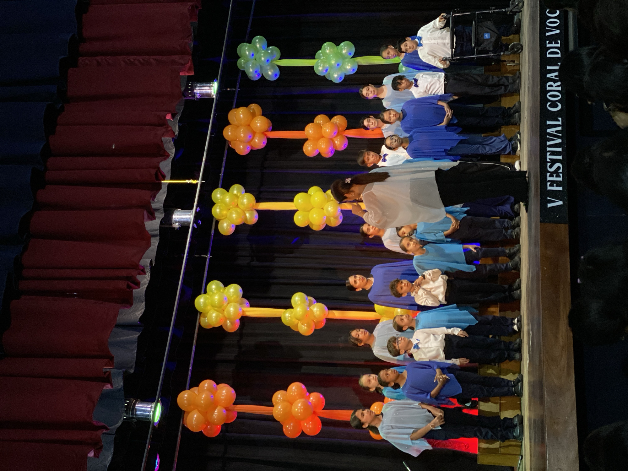 This image captures a vibrant performance or festival event on a stage in Juan Griego, Venezuela, likely indoors during the evening given the stage lighting. A diverse group of children and some adults are arranged in several rows on the stage, facing an audience.
The subjects include a children's choir or performance group. Many children are dressed in various shades of blue tunics or shirts, paired with black pants, while others wear white shirts with blue bow ties and black pants. One child, positioned in the back row on the far right, is seated in a black wheelchair, also wearing a white shirt and blue bow tie. In the front row, slightly to the left of center, an adult, possibly a conductor or leader, stands facing the performers, holding a microphone. The participants appear focused, with some looking towards the conductor and others towards the audience.
The stage is adorned with festive decorations. Clusters of internally lit balloons, arranged in sets of two, are strung horizontally across the dark background. These balloon decorations alternate in color from yellow, orange, to green, connected by fabric strips of corresponding colors. Several stage lights are visible on the floor, casting ambient light on the performers and decorations. Dark red and black curtains frame the left side of the stage.
A notable detail is a black sign with white capital letters positioned on the right side of the stage, reading: "V FESTIVAL CORAL DE VOC". This indicates the event is the "Fifth Choral Festival of Voice" or similar. The atmosphere suggests a formal yet celebratory occasion for a community performance.