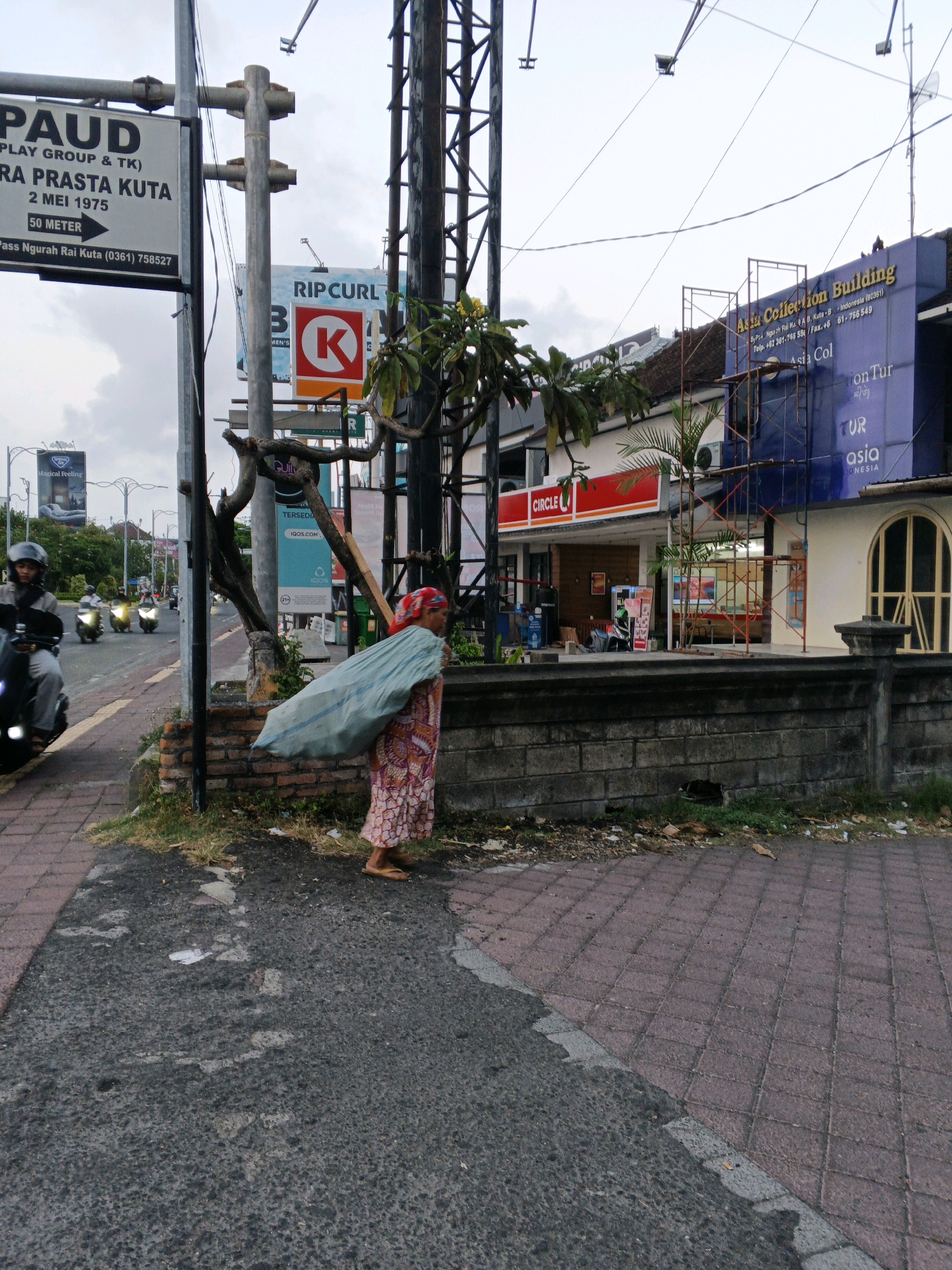 In Kuta, Indonesia, a woman walks along a sidewalk carrying a large, light blue tarp bundle over her right shoulder. She is wearing a floral-patterned dress and a red headscarf. The sidewalk is made of red brick pavers and asphalt. Behind her, a brick wall runs along the edge of a property. Across the street, a Circle K convenience store is visible with a red and white sign. Several motorcycles are passing by on the road, and the sky is overcast. The scene appears to be in a commercial area with various signs and buildings.