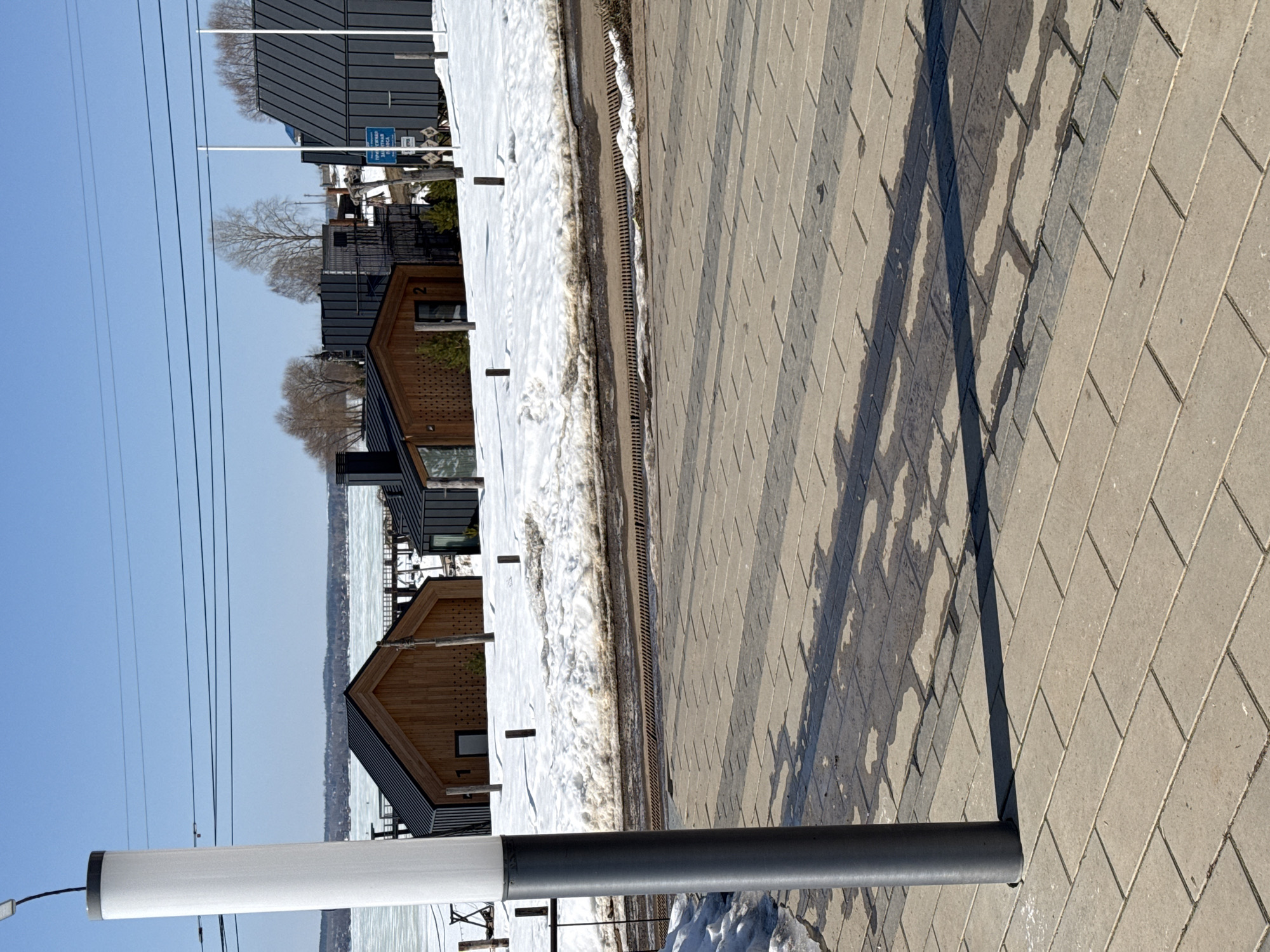 The image depicts a paved walkway in Votkinsk, Russia, adjacent to a waterfront area. The walkway is constructed of light gray rectangular pavers. Shadows cast from unseen objects are visible on the walkway. A row of modern buildings with a mix of wood and dark metal siding is located on the other side of the walkway, separated by a narrow strip of snow-covered ground and a drainage grate. Patches of melting snow are evident, suggesting a transitional season. A light post or similar structure is visible in the foreground, comprised of a white and gray cylindrical post. The sky is clear and blue, with visible power lines. A partially frozen body of water is located beyond the buildings.