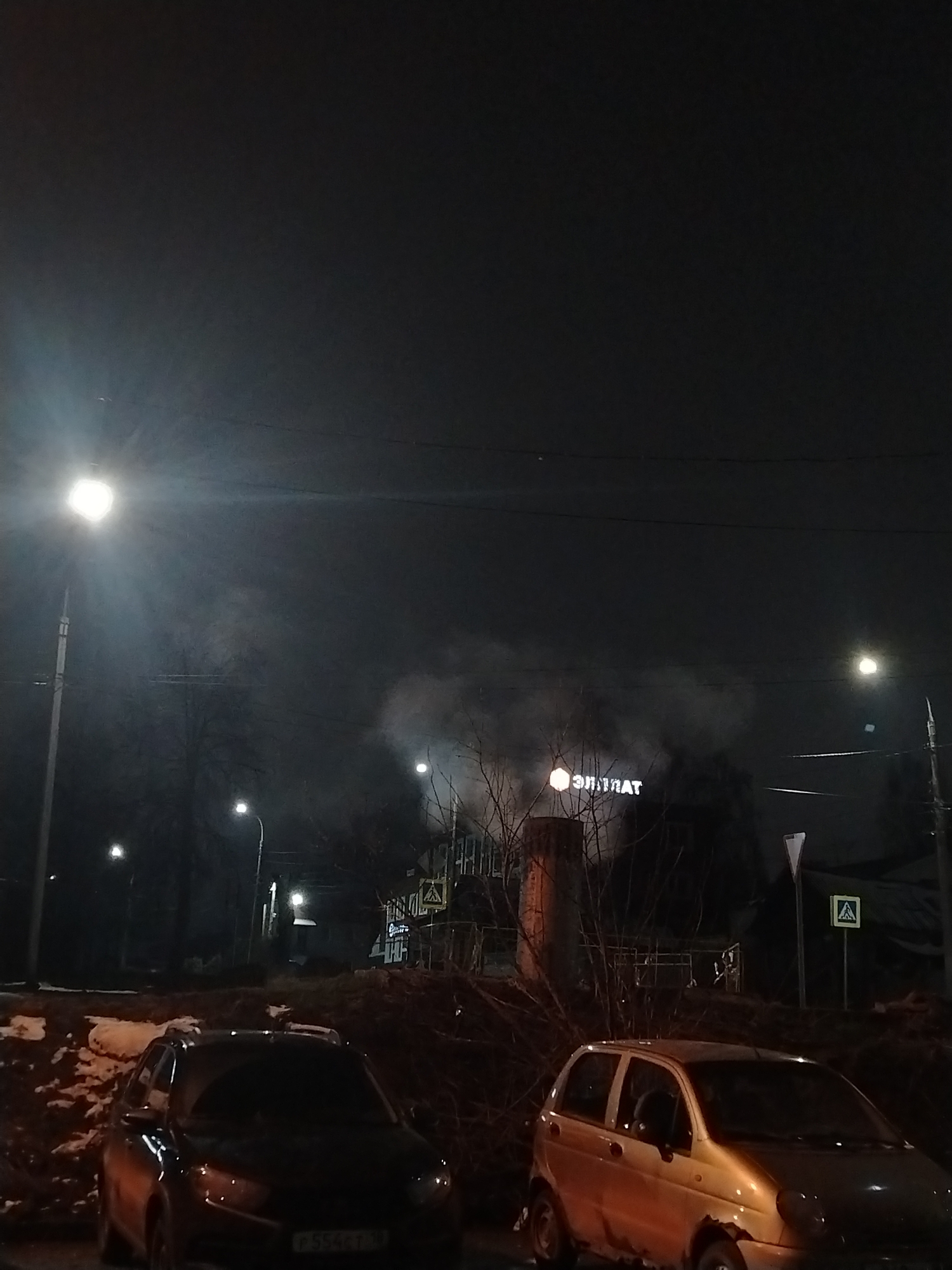 The image shows a nighttime scene in Izhevsk, Russia. In the foreground, two cars are parked: a dark-colored SUV on the left and a light-colored compact car on the right. Behind the cars, there is a slight elevation with bare branches and patches of snow. In the background, a building is visible with a sign that reads "ЭЛИТХАТ" (ELITHAT) illuminated above it. Smoke or vapor is rising behind the building. Streetlights illuminate the scene, and road signs are visible, including a pedestrian crossing sign. The sky is dark.