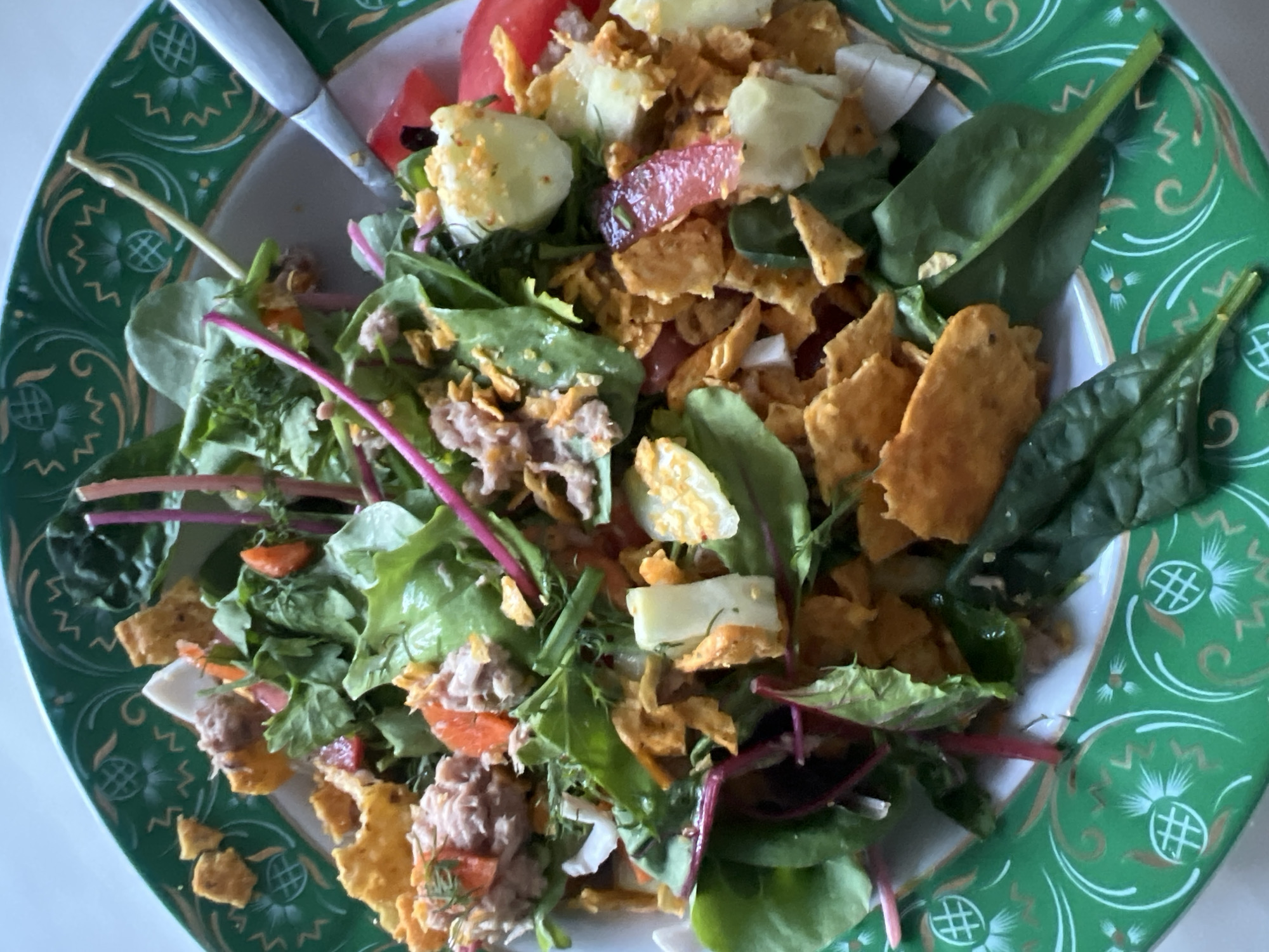 The image shows a close-up view of a salad in a decorative green and white patterned bowl. The salad is composed of various ingredients including leafy greens, tuna, corn chips, and possibly pieces of hard-boiled egg and tomato. A fork or utensil is visible on the right side of the bowl, partially submerged in the salad. The lighting suggests the photo was taken indoors, likely in a domestic setting. There is no indication of the city of Kyiv, Ukraine or any specific location within it.