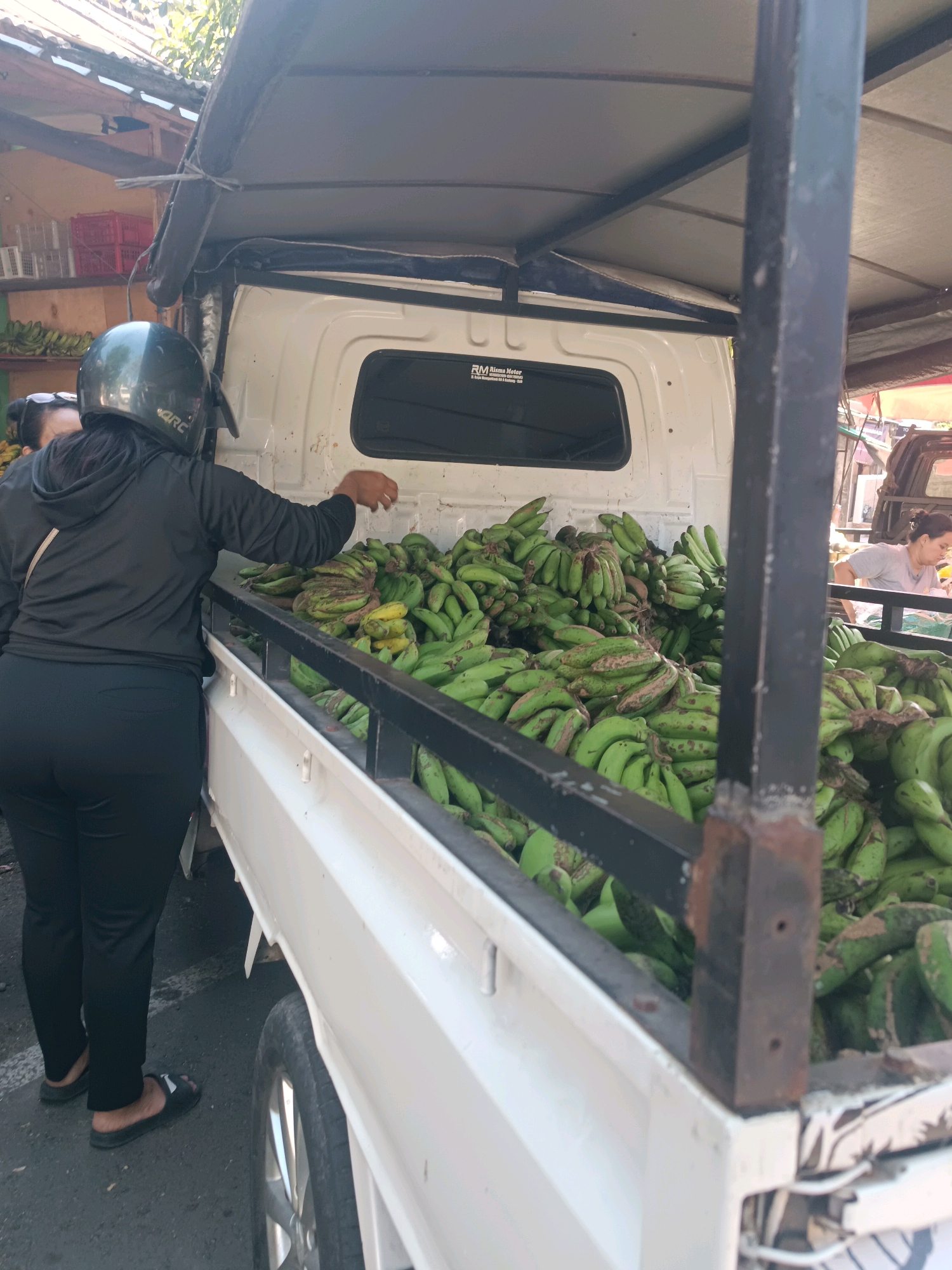In Tabanan, Indonesia, a white truck is parked on a paved street. The truck bed is filled with a large quantity of green bananas. A woman wearing a black hoodie, black pants, and a black helmet is standing next to the truck, reaching into the bed and handling the bananas. Another woman, partially visible, stands behind her. In the background, a vendor is visible behind a table laden with goods, and red and white crates are stacked on shelves. The scene suggests a market or distribution point for fresh produce.