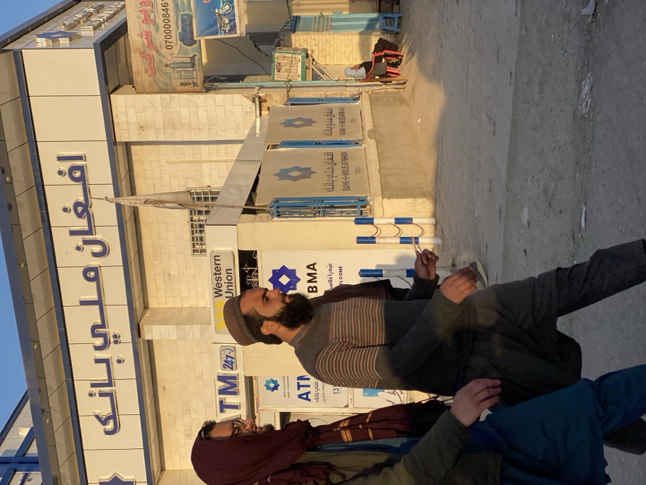 An image taken in Kandahar, Afghanistan, depicts a street scene with a bank and adjacent commercial establishments under clear daylight. The main structure is the Afghan Milli Bank (افغان ملي بانک), identified by blue Pashto/Dari script on a light-colored facade. The bank's ground level features signage for "ATM 24/7," "Western Union," and "BANK-E-MILLIE AFGHAN," along with blue geometric logos. A grey flag, possibly a Taliban flag, is visible near the bank's entrance. Blue and white striped bollards linked by chains demarcate the bank's frontage. To the right of the bank is a narrower commercial unit displaying a sign in Dari/Pashto (partially visible text includes "ویدیو شرکت" and the phone number 0700008461), alongside images of what appear to be gas cylinders and a motorcycle.
In the foreground, two men walk from left to right. The man on the left wears a maroon head covering, glasses, and dark blue trousers (likely part of a salwar kameez). The man in the center wears a dark brown pakol, a grey and brown horizontally striped sweater, dark grey trousers, and has a full dark beard. He holds a small white object in his right hand. Further right, in the midground, a third person wearing dark clothing and a white head covering is seated on a red plastic chair next to a small blue stool. The ground is an unpaved or dusty surface with visible litter. Long shadows are cast across the scene from left to right, indicative of a low sun angle. The sky above is clear blue.