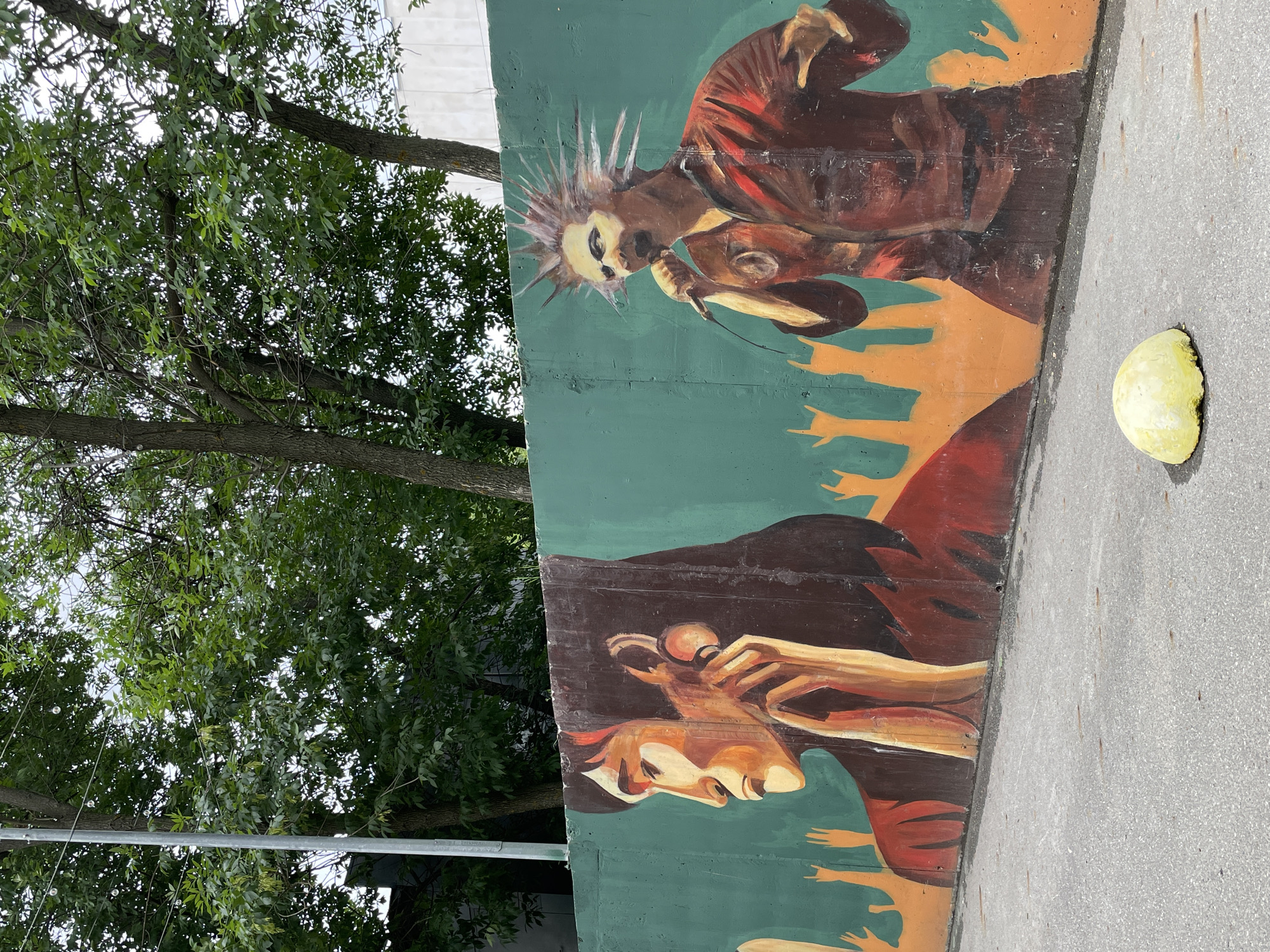 This image shows a mural on a concrete wall in Izhevsk, Russia. The mural features two stylized figures. On the left, a person with long dark hair is depicted in profile, holding a retro-style telephone receiver to their ear. Their facial expression is somber. To the right of this figure is another person with a spiky hairstyle, singing into a microphone. This figure is wearing a red jacket and has an aggressive, energetic expression. Behind the figures, there are abstract representations of raised hands, some forming the "rock on" gesture, suggesting a concert or performance atmosphere. The wall itself is painted a teal green, with an orange-brown base. In the foreground, a small, weathered yellow concrete bollard sits on an asphalt surface. Trees with green foliage are visible above the wall, and a grey pole with string lights runs vertically on the left side of the frame. The sky is overcast.