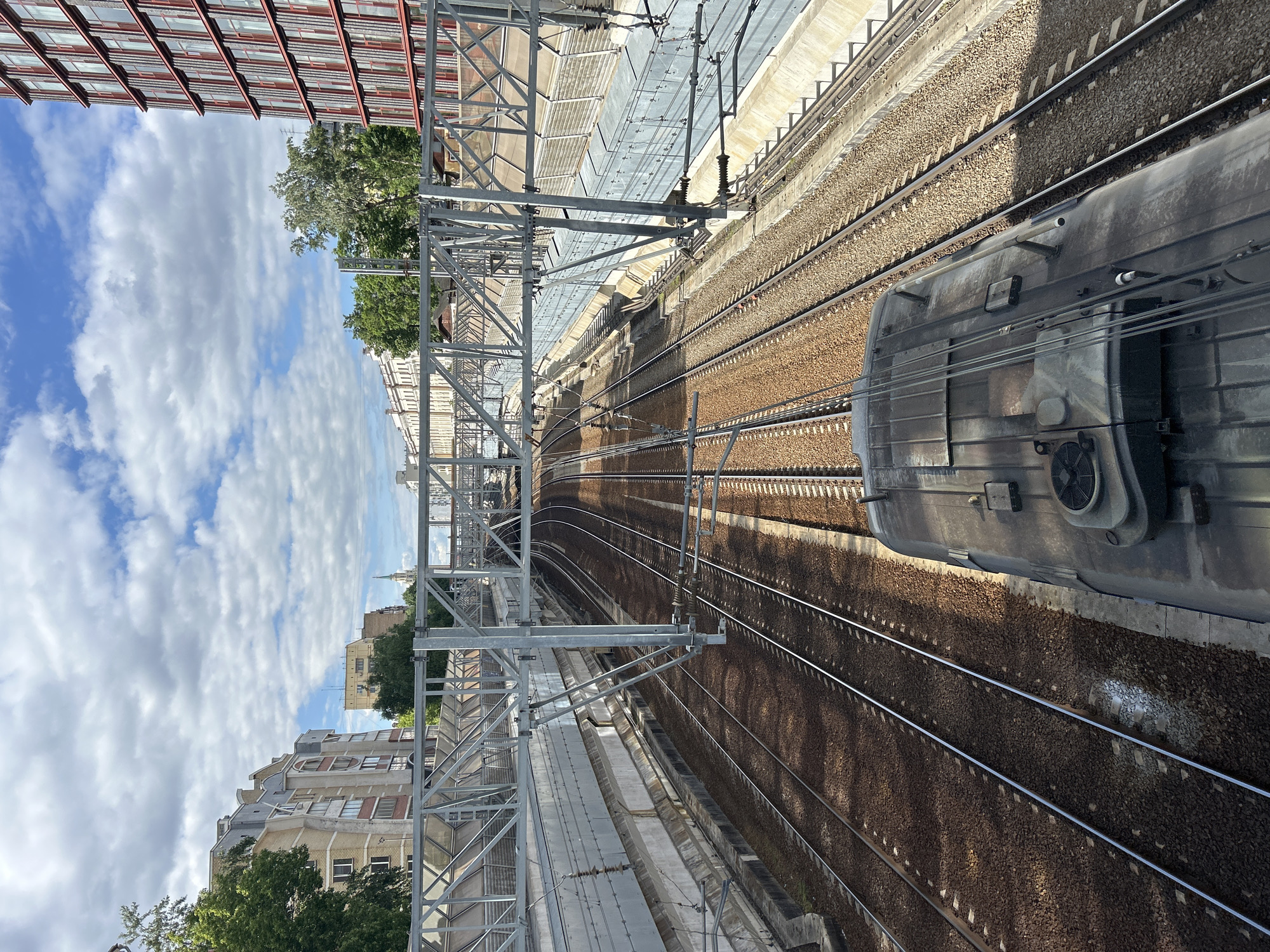 The image presents a view of a railway track, captured from an elevated perspective, likely an overpass or building. The tracks, brown and lined with gravel, curve slightly as they extend into the distance. Overhead electrical lines and metal support structures are prominent on the left side of the frame, providing power to the trains. Part of a train carriage is visible on the right, its exterior appearing metallic and somewhat reflective.
In the background, buildings and trees suggest an urban setting. The sky is visible, filled with white, puffy clouds, providing a bright contrast to the industrial elements of the scene. No people are visible in the image. The location is in the city of Unknown, Unknown.