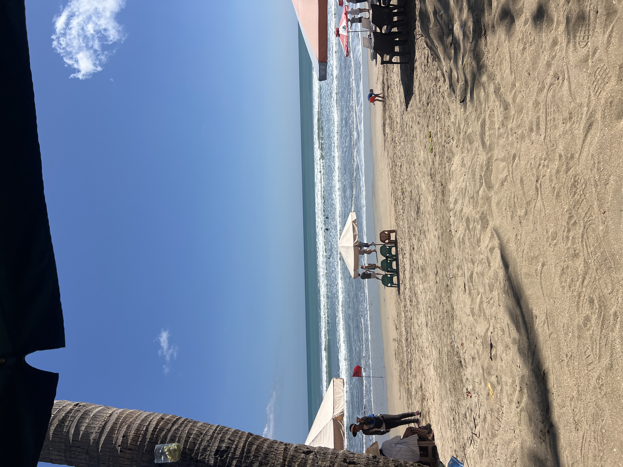 The image depicts a beach scene in Kuta, Indonesia, under a clear blue sky with scattered clouds. The foreground is dominated by sandy beach with shadows cast by unseen objects, possibly palm trees. In the midground, several beach umbrellas are set up, providing shade. Two large, light-colored beach umbrellas are visible, one on the left and another more prominently on the right, with a smaller red and white umbrella between them. Several plastic chairs, some green and some brown, are arranged around the umbrellas.
People are scattered across the beach. On the left, two individuals are seated, one appears to be wearing a straw hat and a striped shirt, while the other, standing, wears a dark jacket and a hat. Near the center, a group of four people are gathered around one of the umbrellas. Further to the right, a child in an orange outfit is seen digging in the sand. A few other individuals are visible walking or standing near the water's edge.
The ocean occupies the background, with moderate waves breaking on the shore. Several people are in the water, some appearing to be swimming or surfing. A red flag is planted in the sand near the water on the left side of the frame, possibly indicating swimming conditions. The overall atmosphere suggests a sunny day at a popular tourist destination.