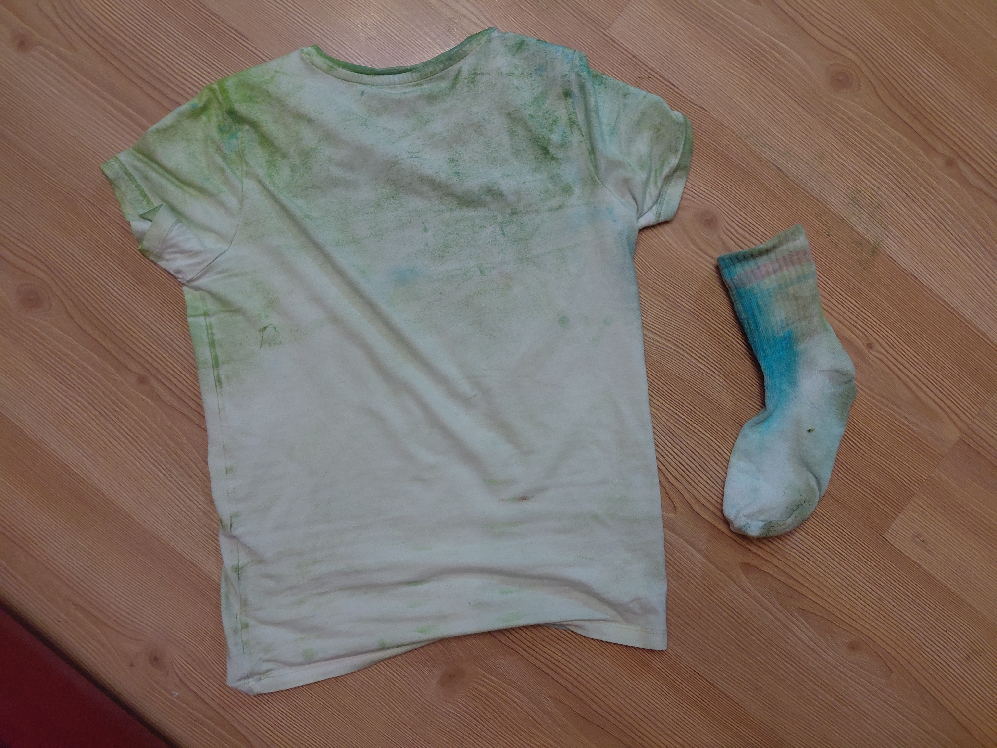 The image displays an indoor scene featuring two items of clothing positioned on a light brown wooden floor. A light-colored, short-sleeved t-shirt occupies the majority of the upper frame, oriented diagonally from the upper left to the lower right. The t-shirt exhibits extensive, irregular staining or dye patterns in shades of green, with some faint blue coloration interspersed, particularly concentrated on the upper right side near the shoulder and collar. The fabric shows minor creasing. Below and to the right of the t-shirt, a single sock is visible, predominantly white with significant patches of blue and green dye, and a subtle pink hue near its cuff. The sock is oriented horizontally. The wooden floor beneath both garments features discernible linear grain patterns and narrow planks running diagonally across the frame. A small portion of a dark red object is visible in the upper left corner of the image. The visual evidence within this image does not provide information to confirm a specific geographical location within the city of Pleven, Bulgaria.