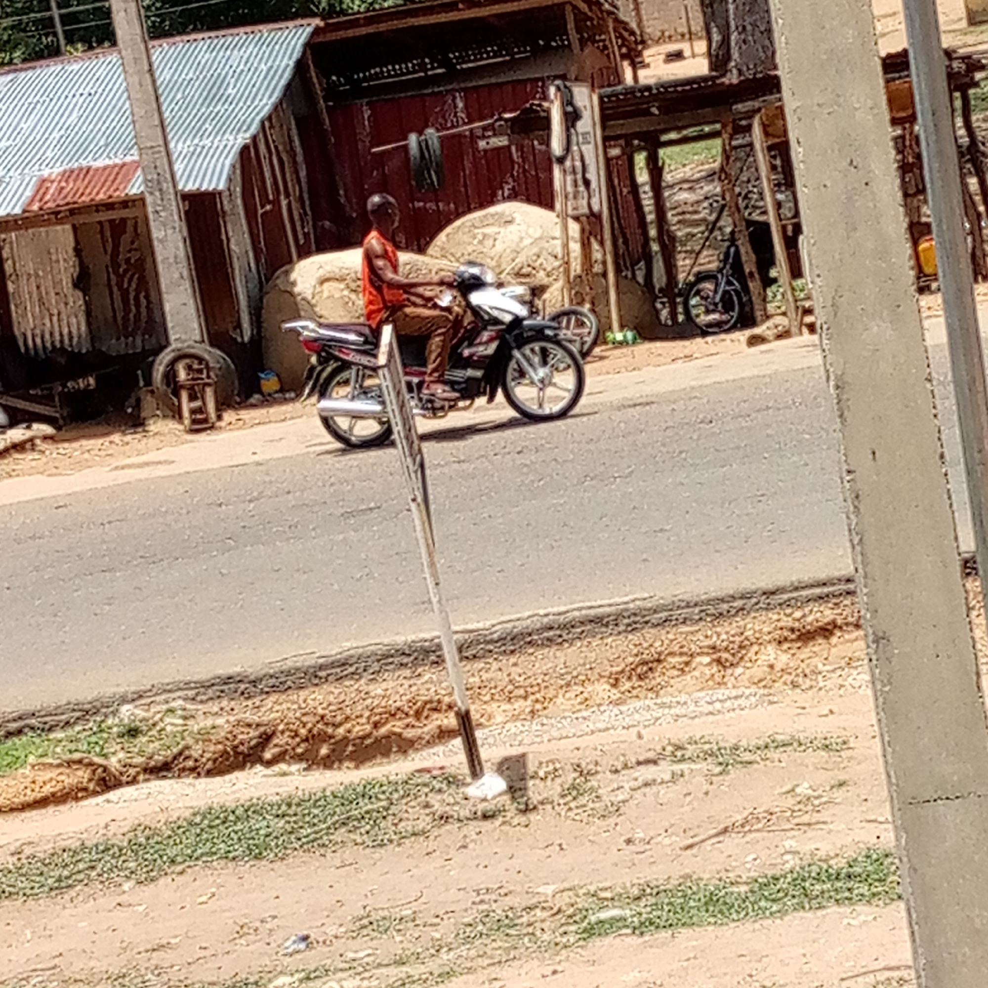 This image depicts a daytime street scene in Mararaba, Nigeria, under bright sunlight.
The main subject is a man of dark complexion, wearing an orange sleeveless shirt and brown trousers, seated on a black and silver motorcycle. He is positioned on a paved road, holding the handlebars, indicating he is either riding or preparing to ride. The motorcycle features silver wheel rims and an exhaust pipe.
In the background, several buildings line the roadside. These structures appear simple and are largely constructed from corrugated metal sheets, some exhibiting rusty red tones while others are silver or grey. Two large, light-colored, irregular objects, possibly sacks or bales, are situated behind the man and his motorcycle, near these buildings. Further back on the right, a bicycle is visible, leaning against a structure, and two tires hang from a bar on one of the buildings. A yellow jerrycan is also present near a building on the far right.
The road is paved and appears light grey, with visible cracks and dirt along its surface. The ground bordering the road is dry, dusty earth with scattered patches of sparse green vegetation. Two prominent, light-colored concrete-like poles stand in the foreground on the right, with a thinner pole positioned closer to the center, slightly obscuring the motorcycle. No visible text is discernible in the image.