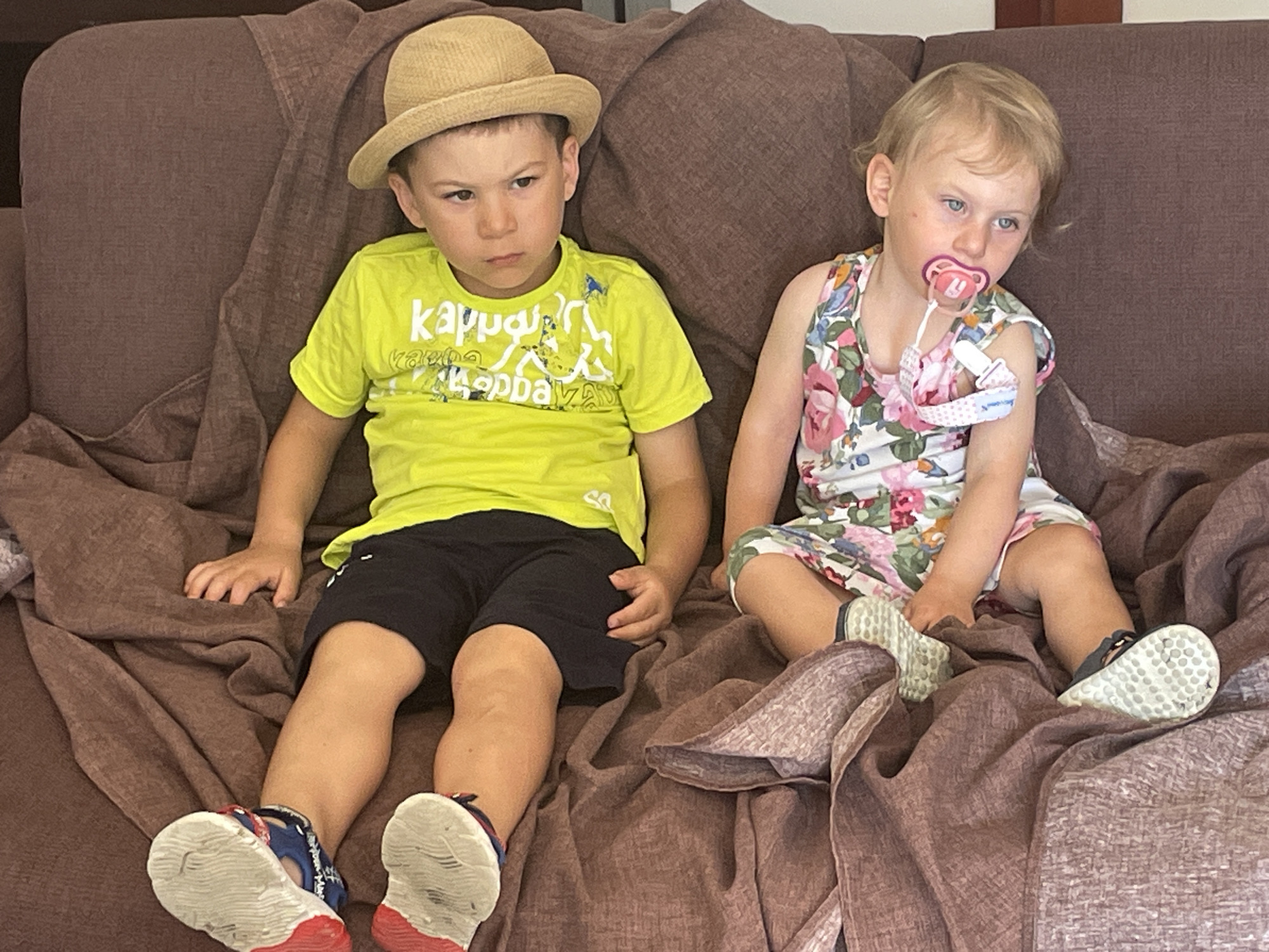Two young children are sitting on a brown couch with a brown blanket draped over it. The child on the left is a boy wearing a straw hat, a neon yellow t-shirt with white lettering, and black shorts. He is looking to his left with a grumpy expression. The child on the right is a younger girl wearing a floral-patterned dress with a pink pacifier in her mouth. She is looking to her right. Both children are wearing sandals. The setting appears to be a living room. The image does not contain enough information to determine the exact location in Punta Ala, Italy.