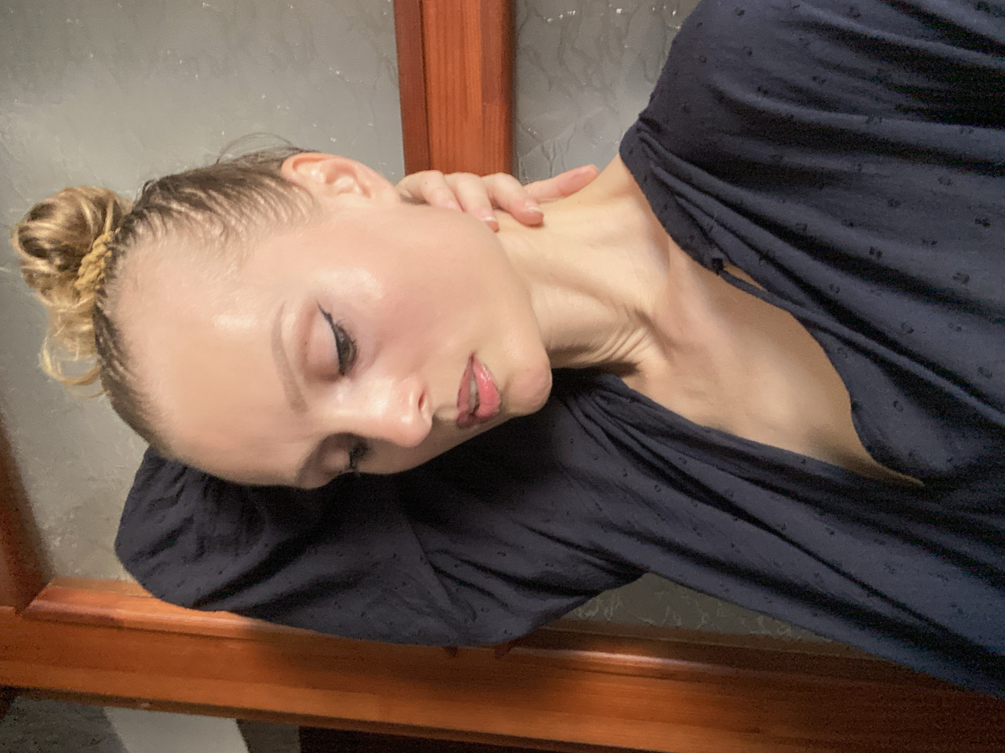 A young Caucasian woman with blonde hair pulled into a bun is shown in a medium, head-and-shoulders shot from the chest up. She is wearing a dark blue top with small, dark polka dots. Her left arm is bent, with her hand resting on the back of her head. Her right hand is touching her neck. She is looking down and to her left. The background appears to be indoors, with a wooden doorframe on the left and a textured wall on the right. No specific location in the city of Lida, Belarus can be identified.