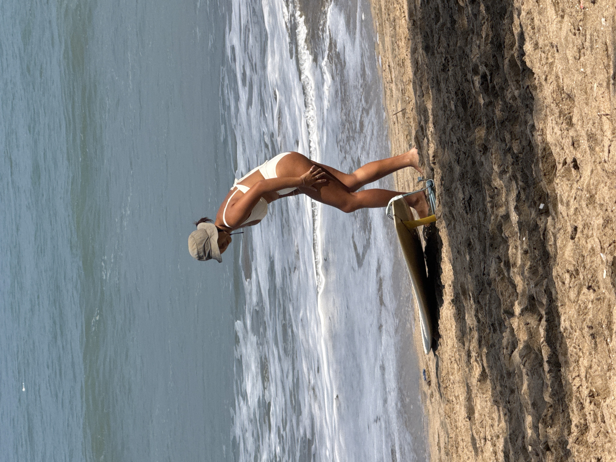 A woman in a white bikini and a beige sun hat stands on a sandy beach in Kuta, Indonesia, bending over a surfboard. The surfboard rests on a blue and yellow stand. The ocean waves are visible behind her, with foam washing ashore. The sand in the foreground is textured, with shadows cast from an unseen object.
