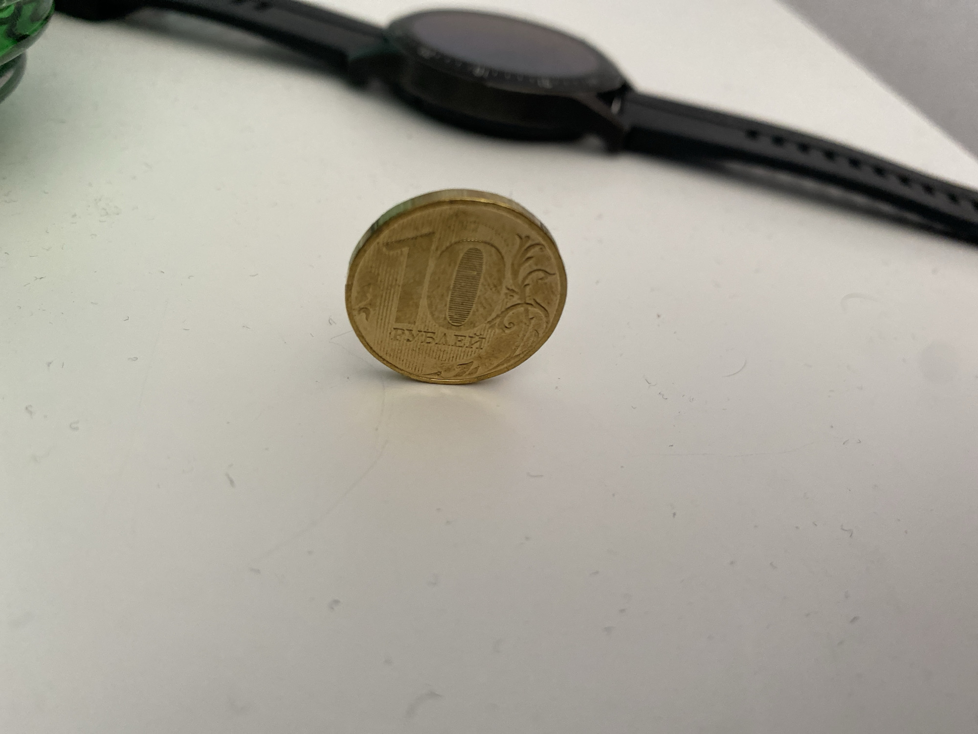 A close-up shot shows a 10-ruble coin, appearing to be of Russian origin, resting on a white surface. The coin is positioned in the foreground, slightly off-center to the left. In the background, out of focus, is the strap and face of a black smartwatch. The environment is a plain, uncluttered surface. There are no people present in the image, and no specific location within Nalchik, Russia, can be identified from the visual information provided.