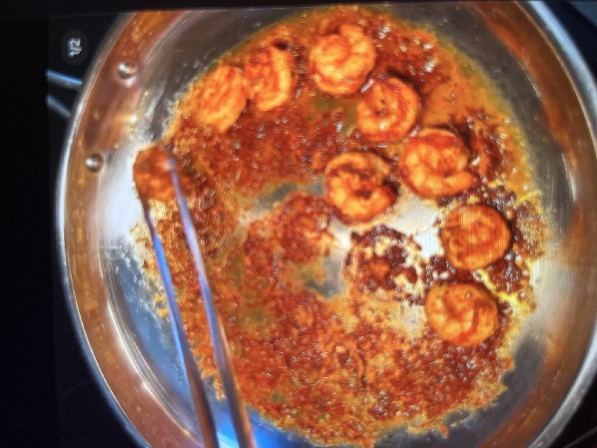 A close-up shot shows shrimp being cooked in a pan. The shrimp are seasoned with a reddish-orange spice mix and are sizzling in a sauce. Tongs are visible, holding some of the shrimp. The pan is metallic and appears to be on a stovetop. The background is dark and out of focus. There is no discernible information within the image to indicate the location of this scene in Abuja, Nigeria.