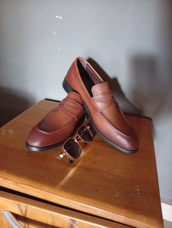 A pair of brown leather loafers rest on a wooden surface, overlapping slightly. A pair of sunglasses with gold frames is positioned next to the shoes. The backdrop is a gray wall. The lighting is soft, casting shadows that enhance the shape of the items. The composition is simple, focusing on the texture and color of the shoes and sunglasses.