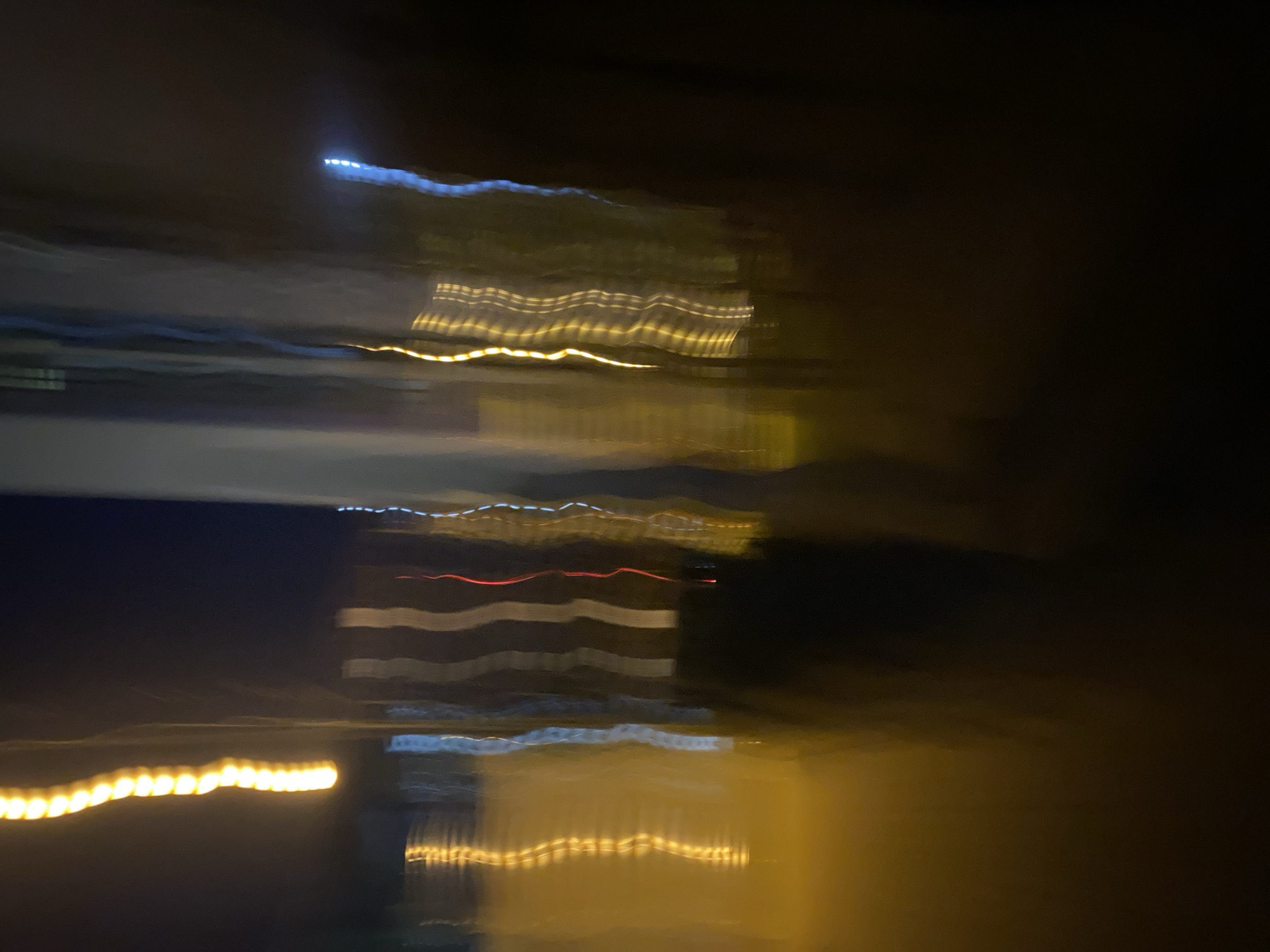 The image is an abstract, blurred depiction of city lights at night, likely from a moving vehicle in Kyiv, Ukraine. Streaks of yellow and white light dominate the scene, suggesting streetlights or illuminated building windows. Vertical lines of blue and red light are also visible, possibly from traffic signals or vehicle taillights. The overall impression is one of movement and the transient nature of urban nightlife. No specific landmarks or people are clearly identifiable due to the intentional blurring and abstract composition. The dark background indicates nighttime conditions.
