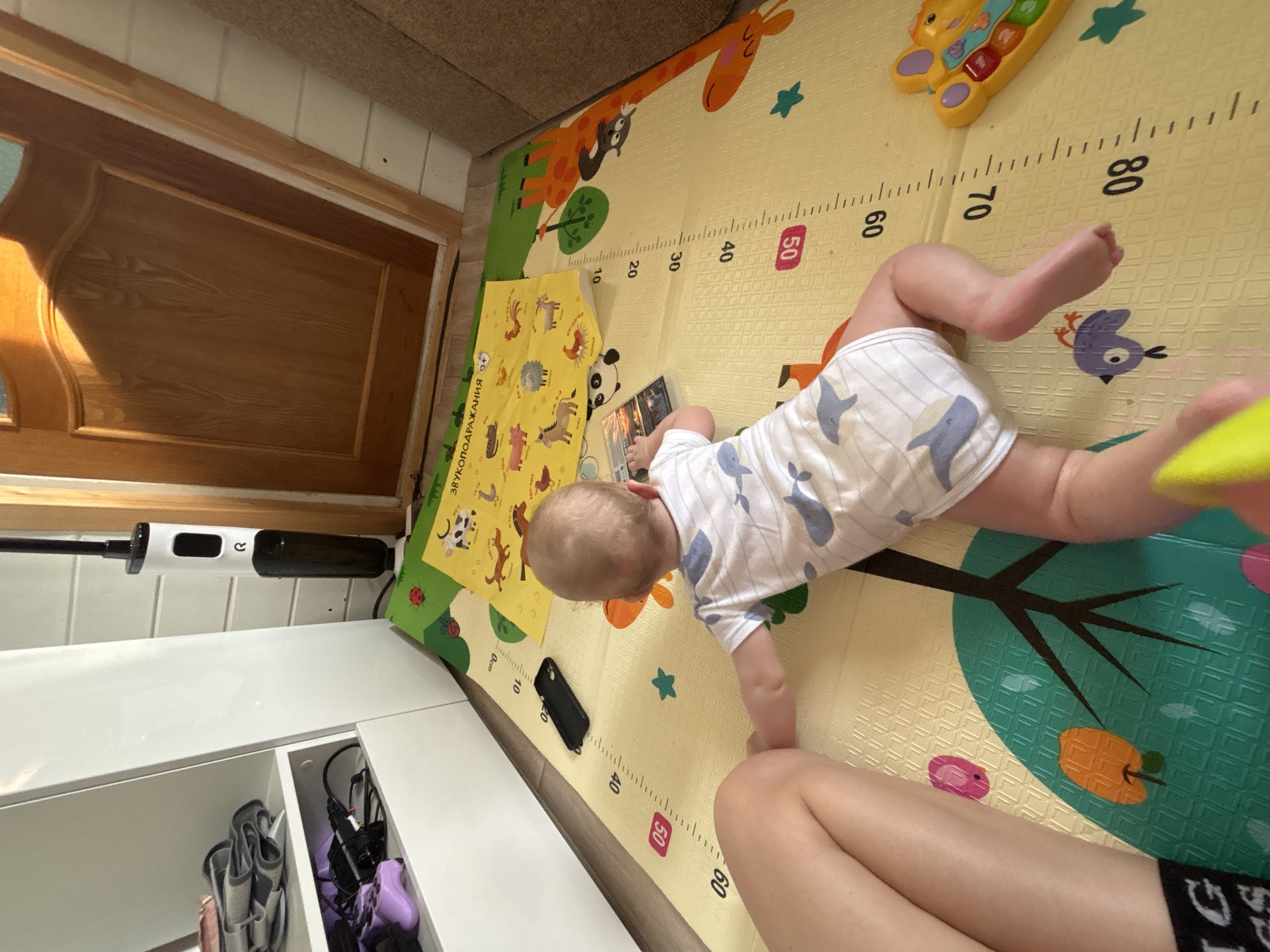 A baby is lying on a play mat in what appears to be a home setting. The baby is wearing a white and blue patterned onesie. The play mat is yellow with green and orange illustrations and measurements marked in centimeters. The baby is reaching for an object held by an adult, whose legs and clothing are visible. A white cabinet and various household items are present in the background. The scene is within a dwelling, location in the city of Раздолье, Russia.