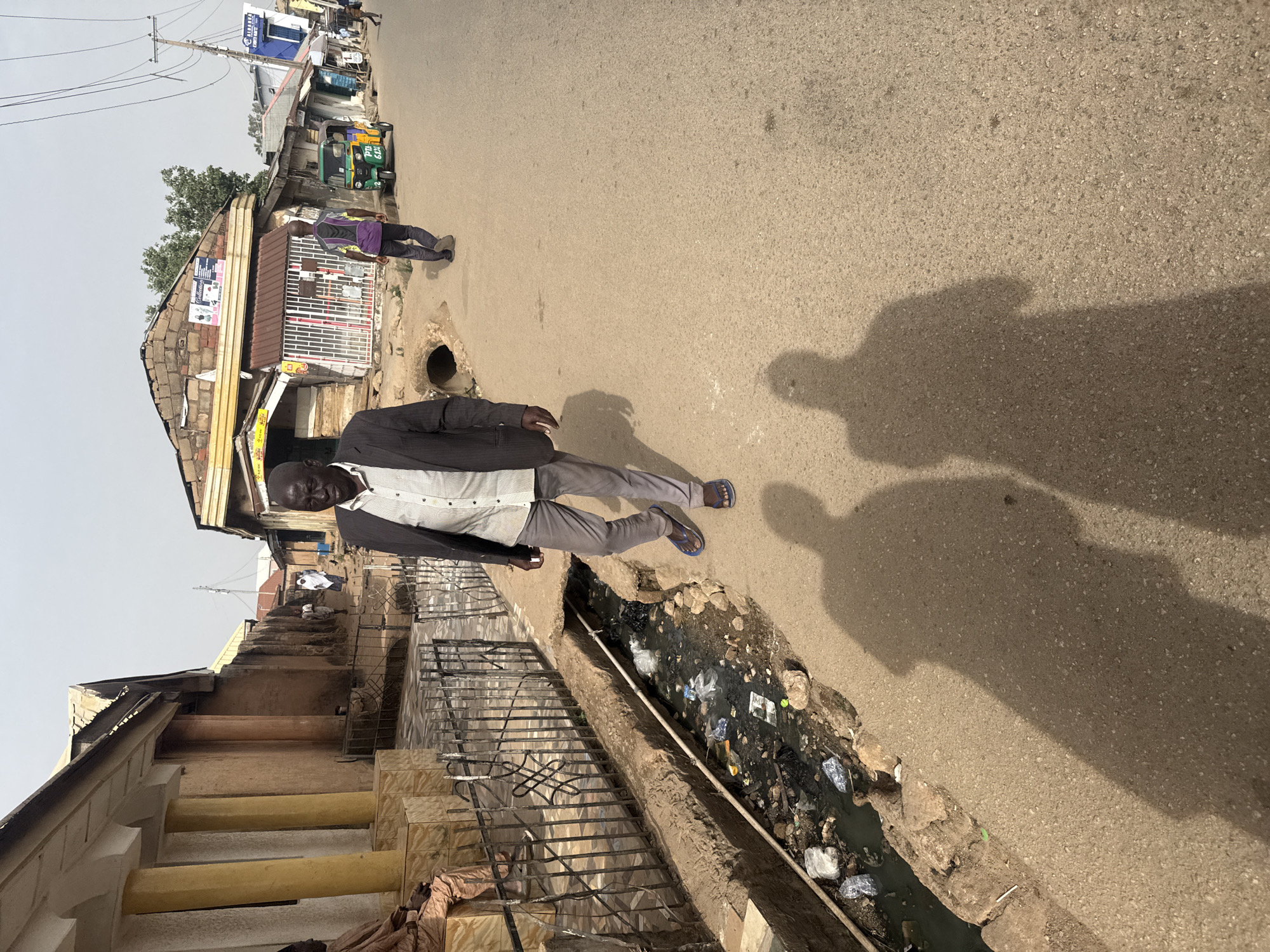 An adult male wearing a black coat, white shirt, and gray pants is walking on a paved road in Jos, Nigeria. The environment includes buildings, a drainage ditch filled with trash, and other people further down the street. Shadows are cast on the road surface.