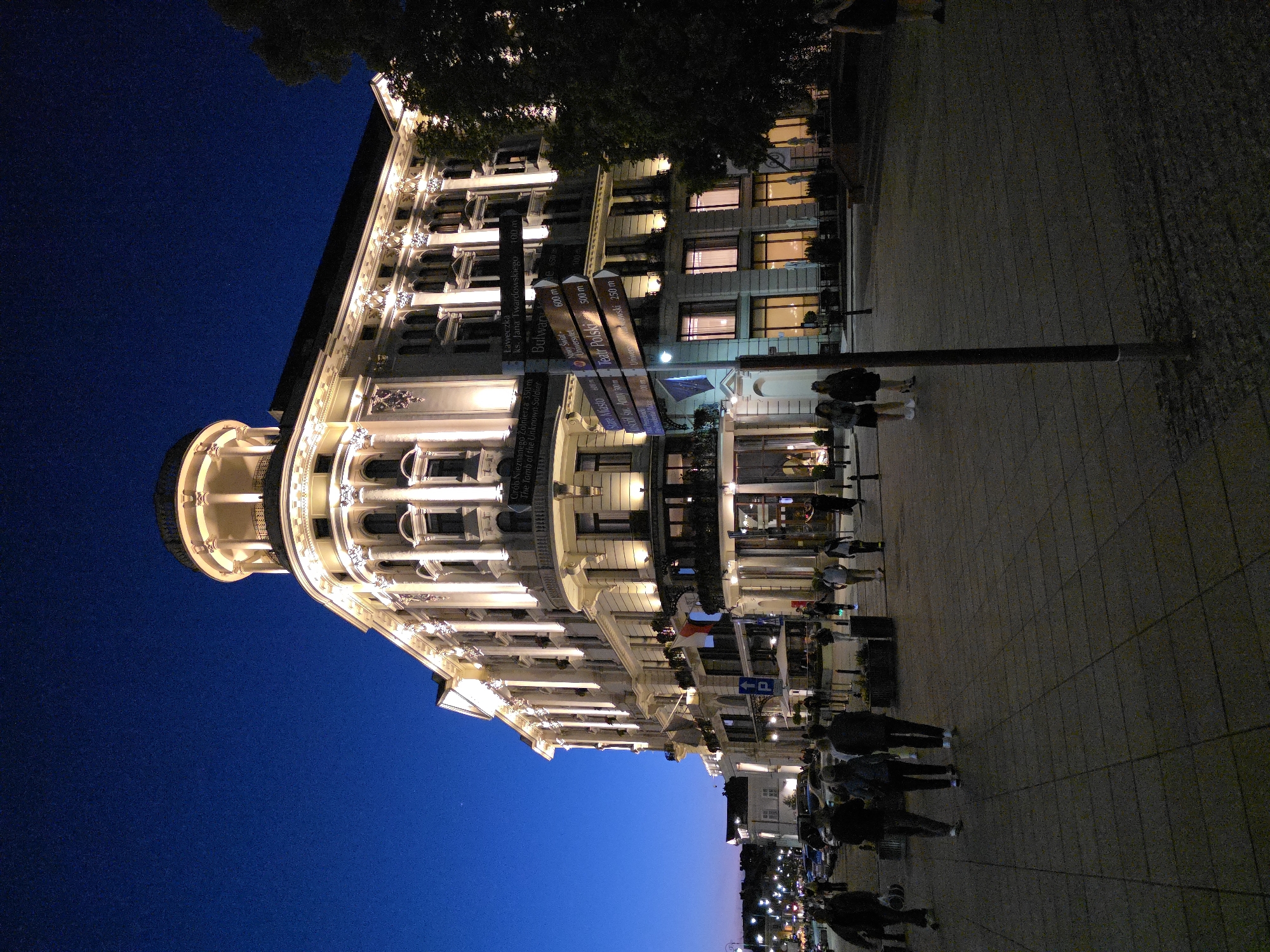 Captured at night, the image presents a view of a street scene in Warsaw, Poland. Dominating the scene is an illuminated building with architectural details, including a rounded section at the top and multiple windows. A signpost is visible, displaying distances to local landmarks such as "Grób Nieznanego Żołnierza" (Tomb of the Unknown Soldier), "Stare Miasto" (Old Town), "Teatr Polski" (Polish Theatre), and "Uniwersytet" (University). Several people are seen walking along the sidewalk in front of the building. The sky is a deep blue, contrasting with the warm lights of the building and street. The ground is paved, and additional lights are visible in the distance. The image is slightly rotated, causing a tilted perspective.