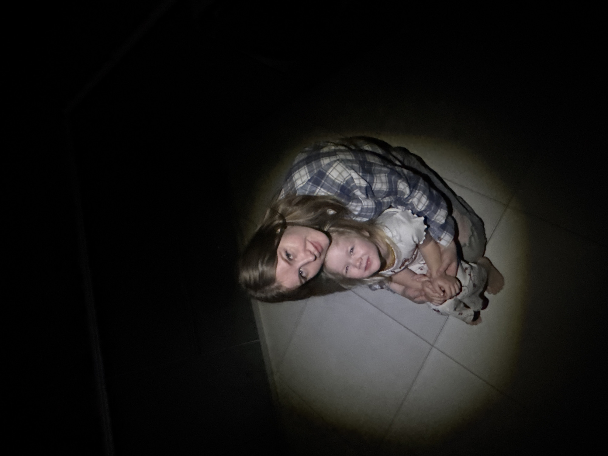 A young woman and a small child are illuminated by a light source, possibly a flashlight, in a dark environment. The woman is wearing a plaid shirt. The child is wearing a light-colored shirt and patterned pants. They are embracing each other on a tiled floor. The surrounding environment is almost completely dark. There is no evidence to suggest this image was taken in Gornyi (Dvurechenskoe S/P), Russia.
