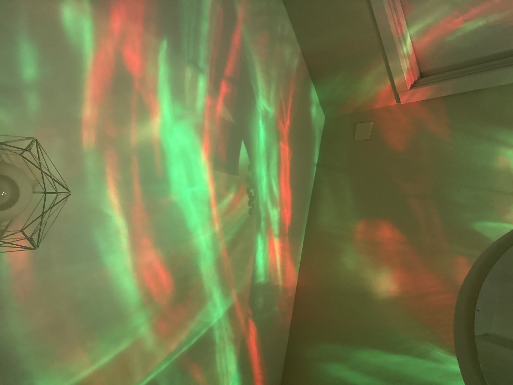The image is an interior shot, likely taken indoors at night. Red and green lights are projected onto the walls, creating an abstract pattern. A modern chandelier with a geometric design is visible in the upper left corner. A window frame is present in the upper right. A round object is on the bottom right corner. The overall environment appears to be a room with neutral-colored walls, suggesting a residential or commercial space. There is no information to suggest the location is Batumi, Georgia.