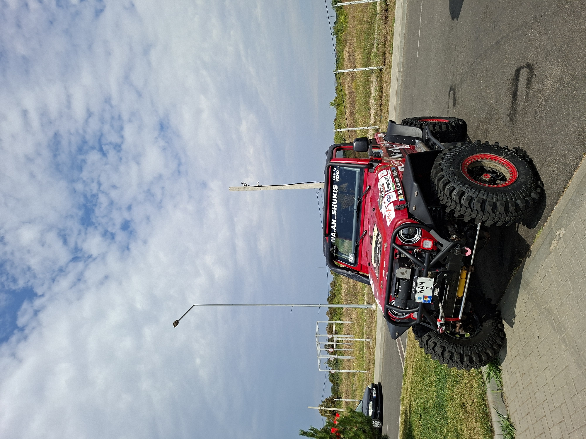 The image depicts a custom red off-road vehicle, likely a Jeep Wrangler, parked on an asphalt road adjacent to a paved walkway and a grassy area. The vehicle is heavily modified with large, aggressive tires, a prominent roll cage, and numerous decals and stickers on its exterior. The license plate visible reads "NAN 1" with a Moldovan flag emblem.
In the background, a black sedan is visible on the road, and beyond that, a field with sparse vegetation and a white fence. A tall lamp post stands to the left of the off-road vehicle, and a structure resembling a metal frame is visible further in the background. The sky is partly cloudy with patches of blue sky and white clouds. The presence of the Moldovan license plate and the landscape are consistent with a location in or around Stăuceni, Moldova. The scene appears to be an outdoor parking area on a sunny day.